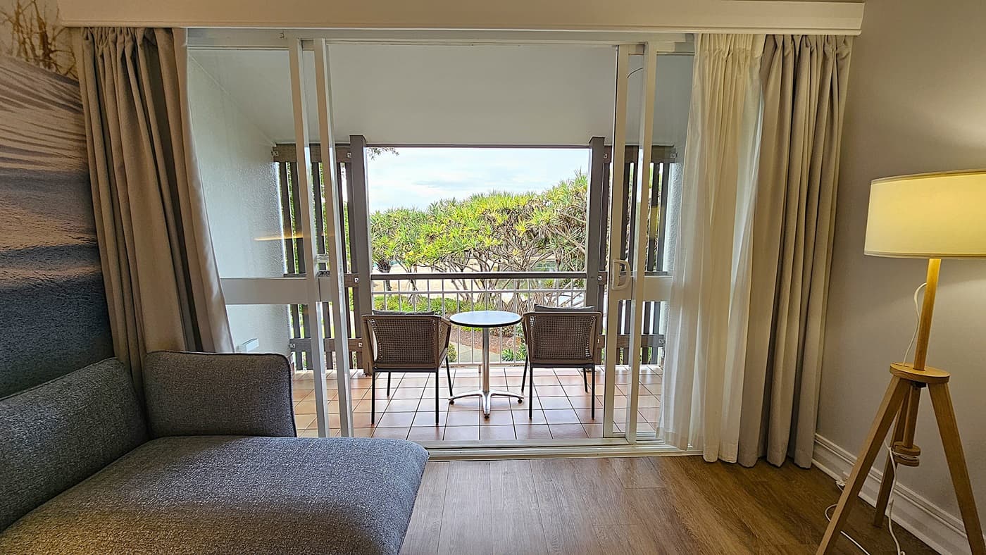 On the left, a grey upholstered chaise lounge with a beach-themed mural on the wall behind it. To the right, a wooden tripod floor lamp with a beige shade. In the middle, a sliding glass door opens to a balcony with two wicker chairs and a round table, overlooking greenery. The floor is wooden inside and tiled on the balcony. The ceiling is white with recessed lighting.