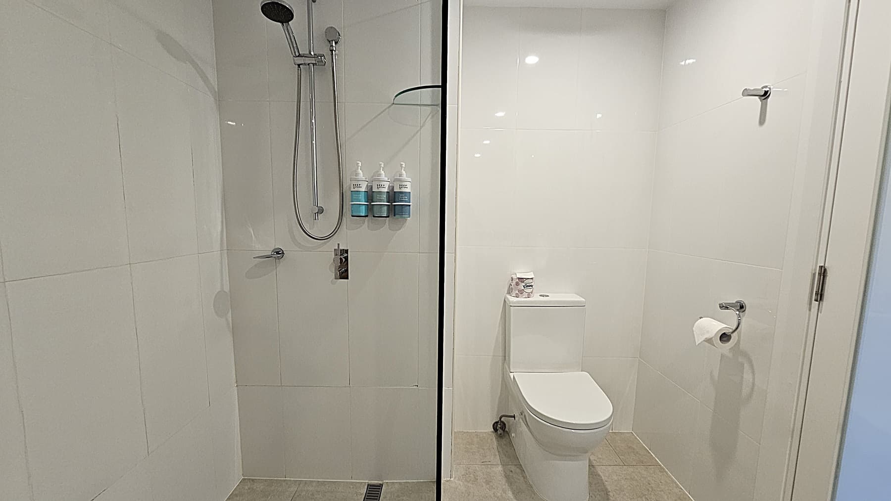 On the left, a shower area with a wall-mounted showerhead, a handheld shower, and three soap dispensers. To the right, a white toilet with a roll of toilet paper on a holder and a small shelf above it holding additional toilet paper rolls. The floor is tiled in light grey, and the ceiling is white with recessed lighting.