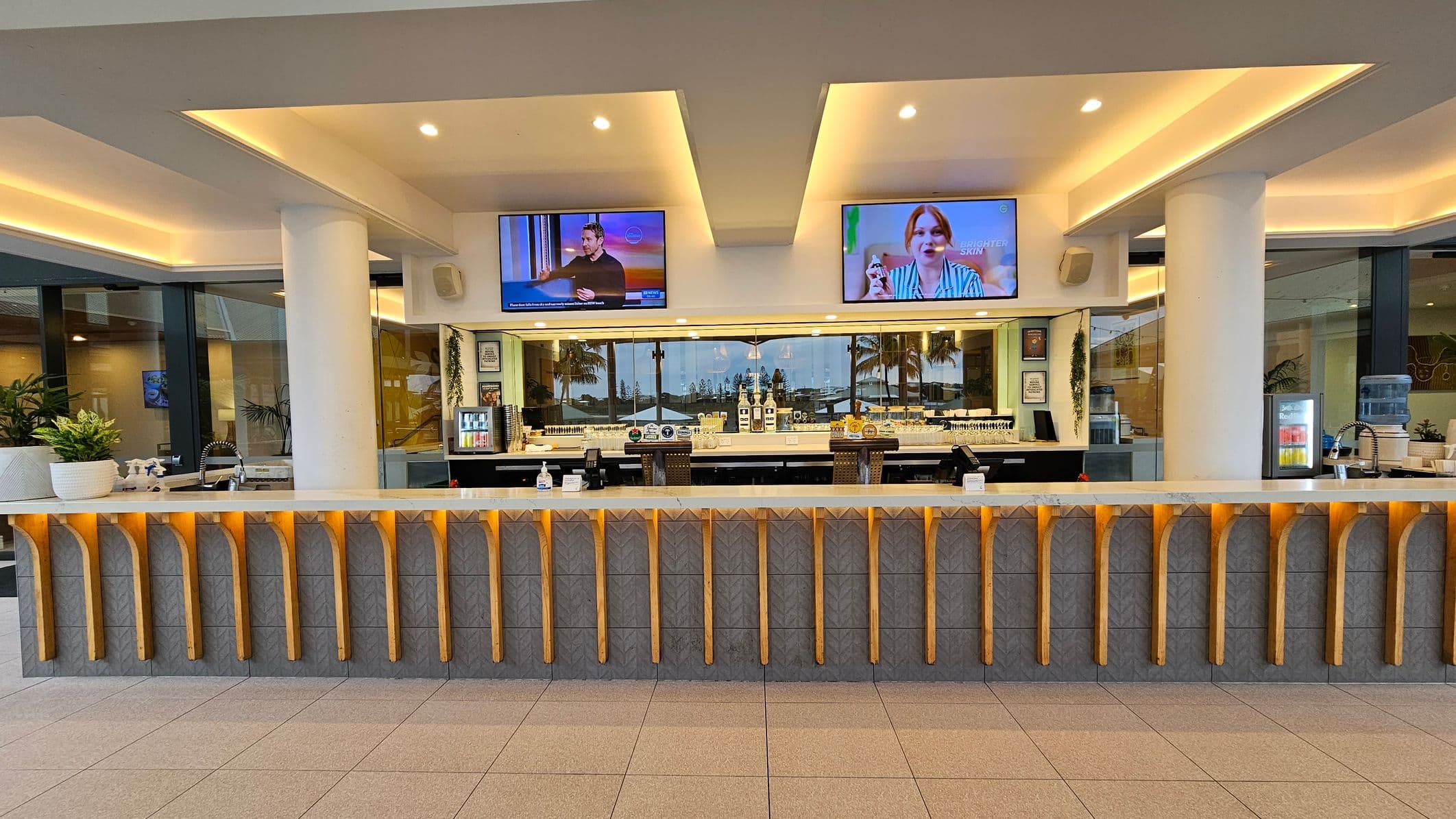 Spacious interior with a high, wooden-beamed ceiling. On the left, a yellow wall with artwork and a staircase with a metal railing. In the centre, a seating area with wooden chairs, tables, and a grey partition. Above, large, decorative pendant lights. On the right, a long counter and a white wall with framed art. The floor is tiled, and there are potted plants near the walls.