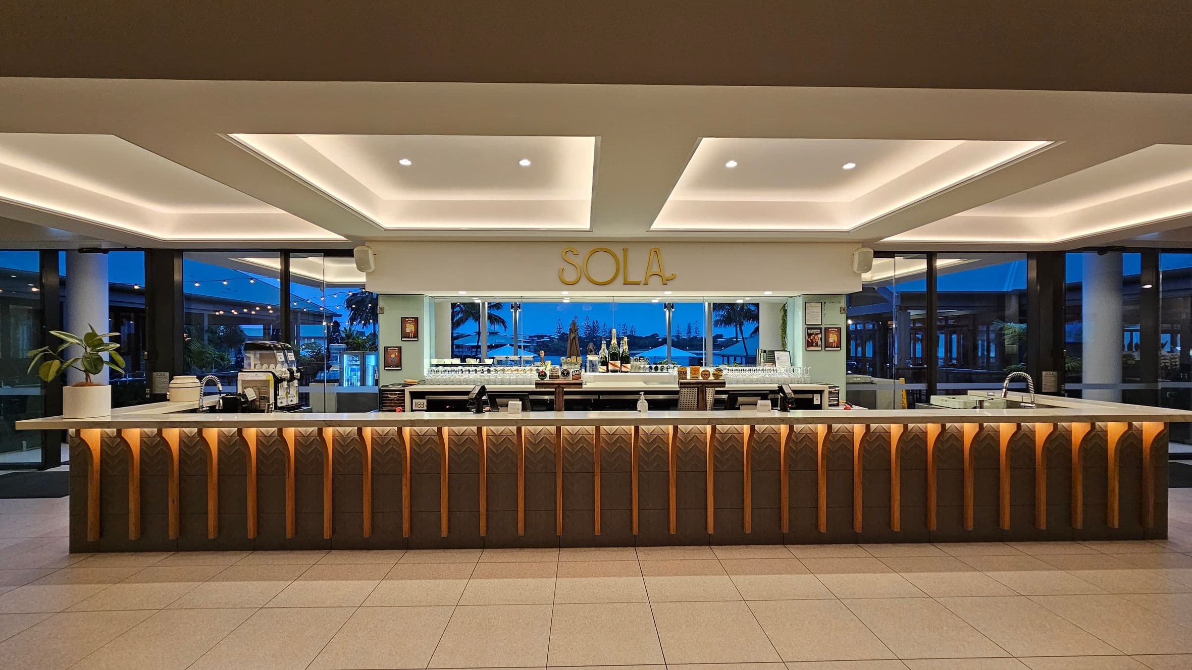 A modern bar area with a long counter featuring geometric patterns and warm lighting. Behind the counter, there are shelves with bottles and glasses, and a sign reading "SOLA" above. Large windows in the background reveal an outdoor view with palm trees and a blue sky. The ceiling has recessed lighting, and the floor is tiled. A plant is placed on the left side of the counter.