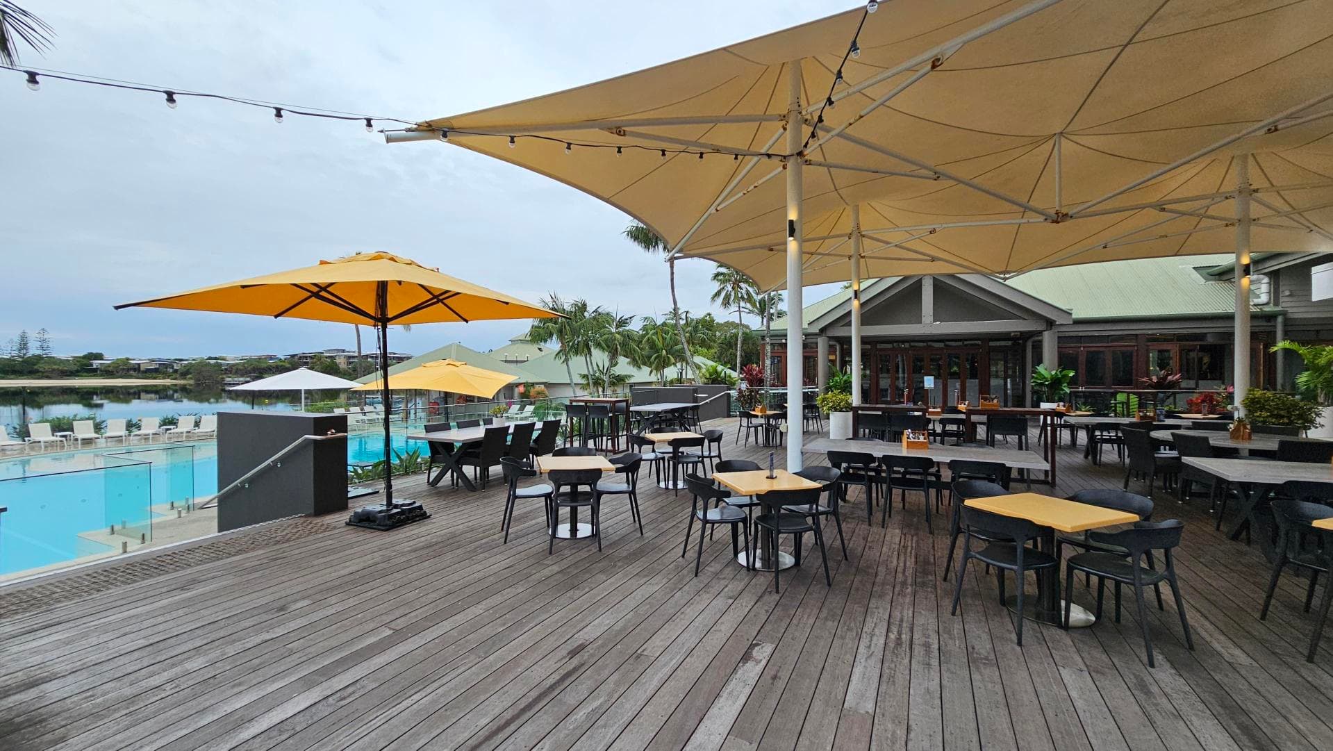 Outdoor dining area with wooden decking and several tables and chairs. Large yellow umbrellas provide shade. In the background, there's a swimming pool with sun loungers and a glass railing. A building with a green roof is visible, surrounded by palm trees and plants. Overhead, string lights are hung across the area.