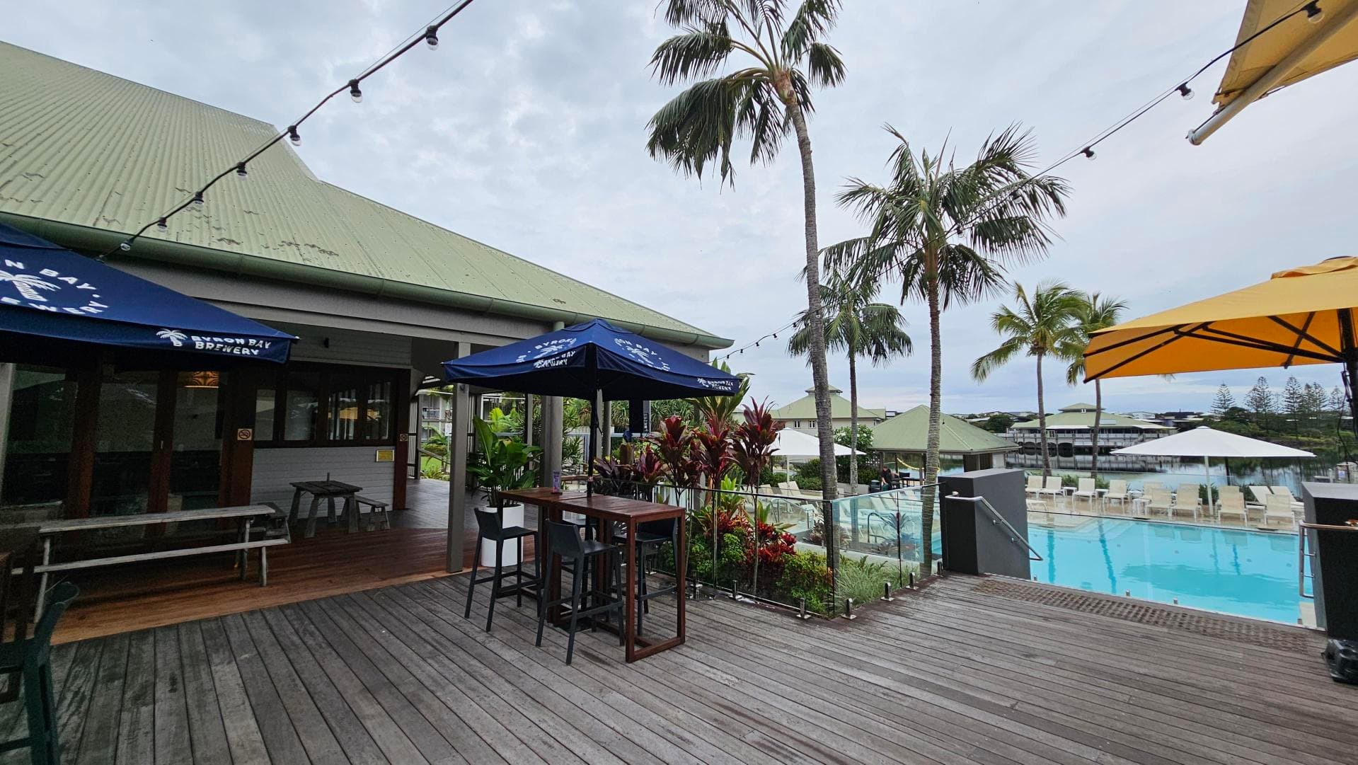 A wooden deck with outdoor seating, including tables and chairs under blue and yellow umbrellas. On the left, there's a building with a green roof and a sign on the umbrella. In the middle, potted plants and palm trees add greenery. On the right, a swimming pool is surrounded by white loungers and umbrellas. String lights are hung above, and the area has a relaxed, open-air atmosphere.
