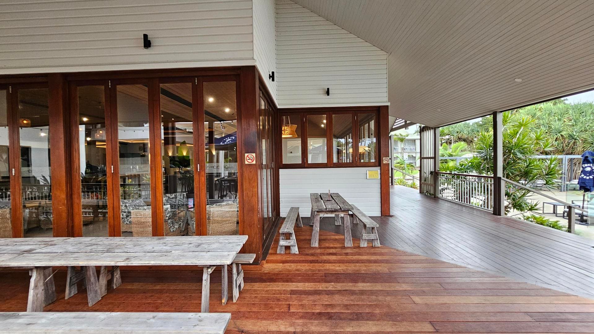 Wooden outdoor seating area with two rustic picnic tables on a wooden deck. On the left, there are large glass doors with wooden frames leading to an indoor space. The ceiling is sloped and covered with light-coloured panels. On the right, a railing borders the deck, with lush green plants visible beyond. The area is sheltered, providing a cosy atmosphere.
