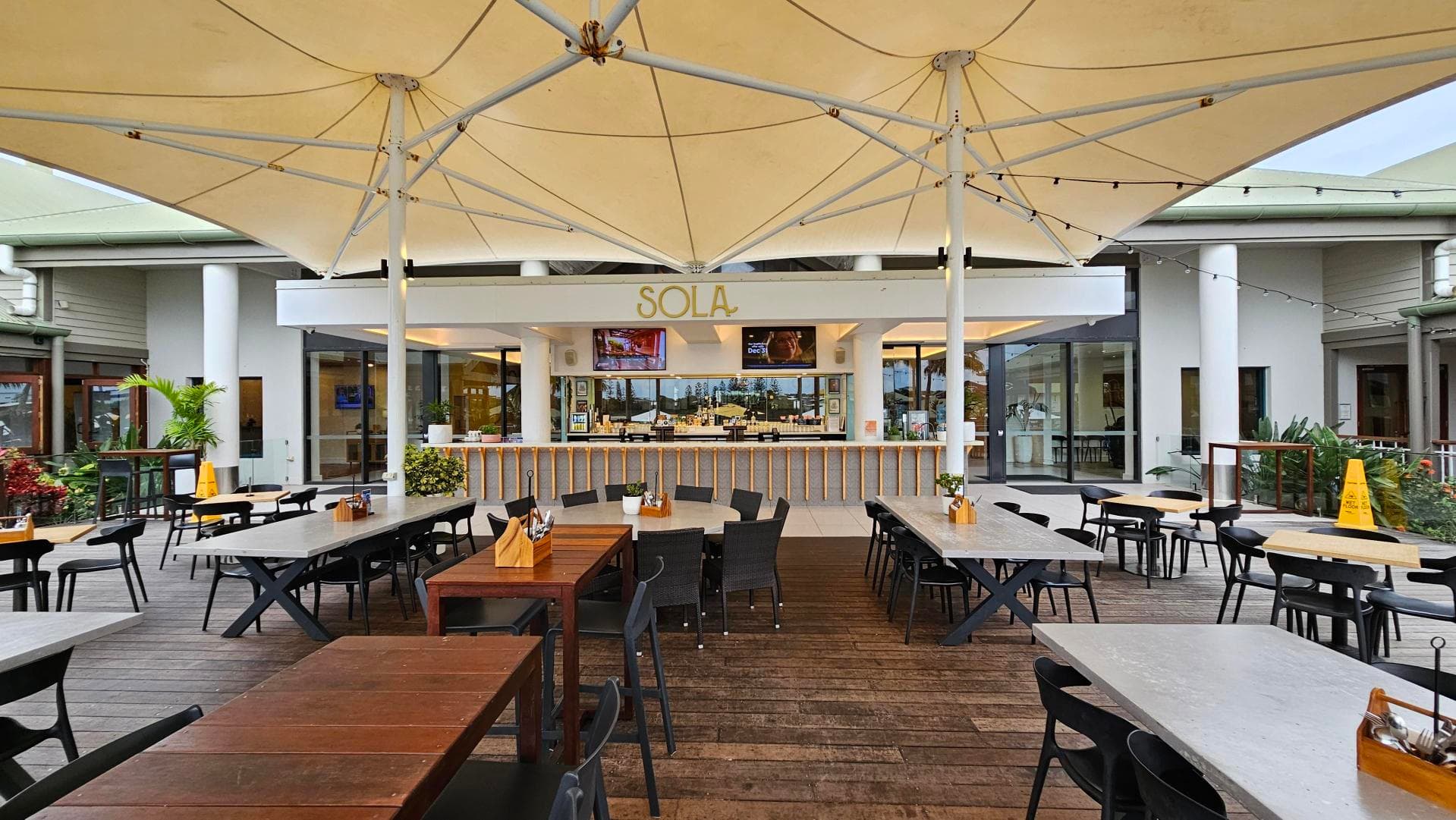 Outdoor dining area with wooden and metal tables and black chairs on a wooden deck. A large white canopy covers the space. In the background, a bar named "SOLA" with a modern design, featuring two large screens and glass doors. Potted plants and a yellow caution sign are visible. The area is surrounded by buildings with white pillars and green roofs.