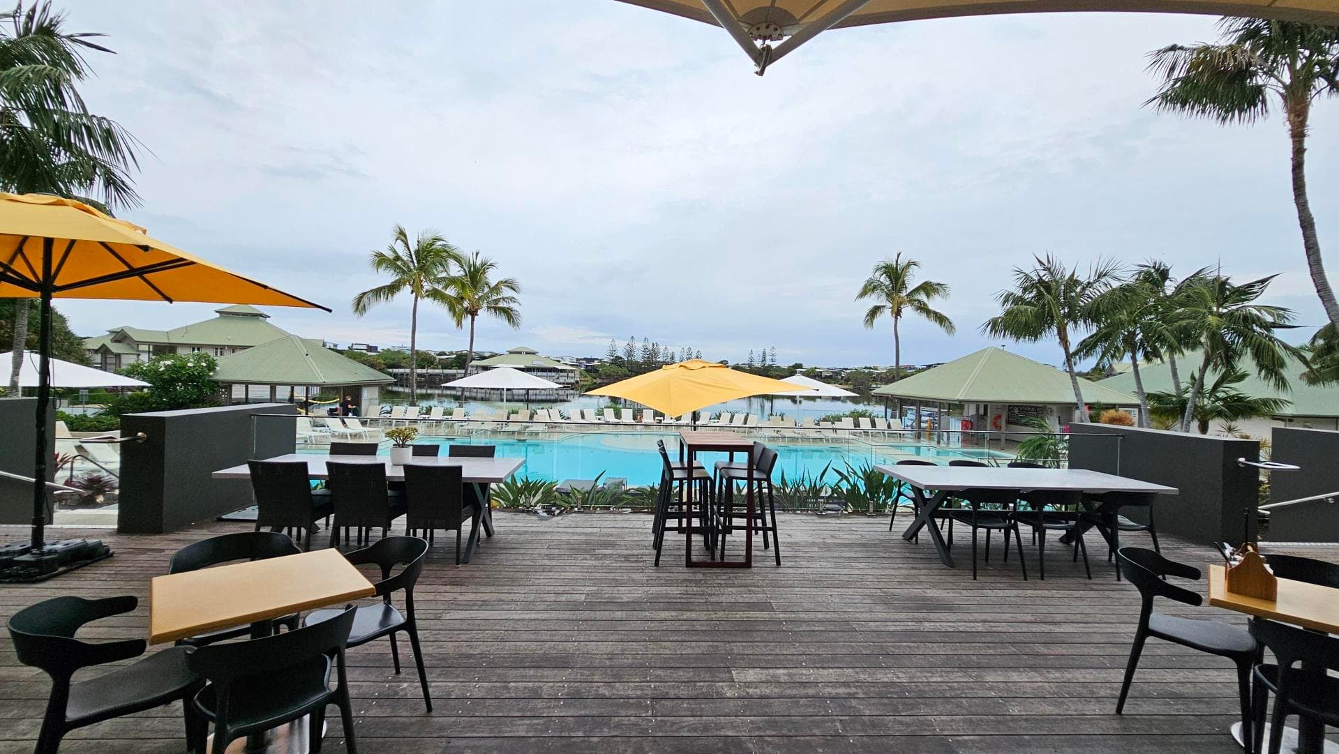 Wooden deck with tables and chairs in the foreground, some under yellow umbrellas. Beyond the deck is a swimming pool surrounded by sun loungers and more umbrellas. In the background, there are palm trees and buildings with green roofs.