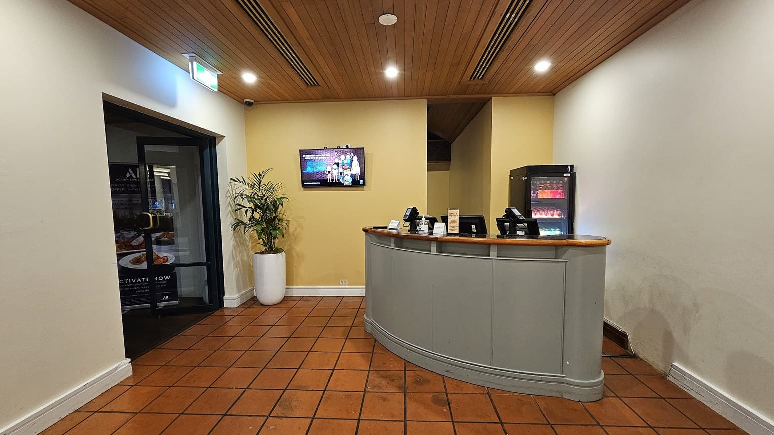 On the left, a black-framed glass door leads to another room, with a large potted plant next to it. To the right, a curved reception desk with a wooden top holds several electronic devices and a small display stand. In the background, a black refrigerator with illuminated shelves is against a yellow wall. The floor is covered in terracotta tiles, and the ceiling is wooden with recessed lighting and ventilation panels.