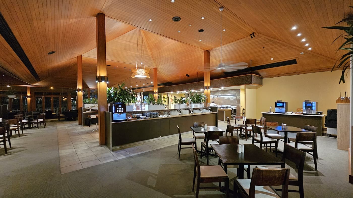 On the left, several dining tables with wooden chairs are arranged on a carpeted floor, with large windows and wooden columns. To the right, more dining tables and chairs are set on a carpeted area, with a beverage station against a yellow wall. In the middle, a long buffet counter with various food items and potted plants is positioned on a tiled floor. The ceiling is wooden with recessed lighting, pendant lights, and ceiling fans, creating a warm and well-lit atmosphere.