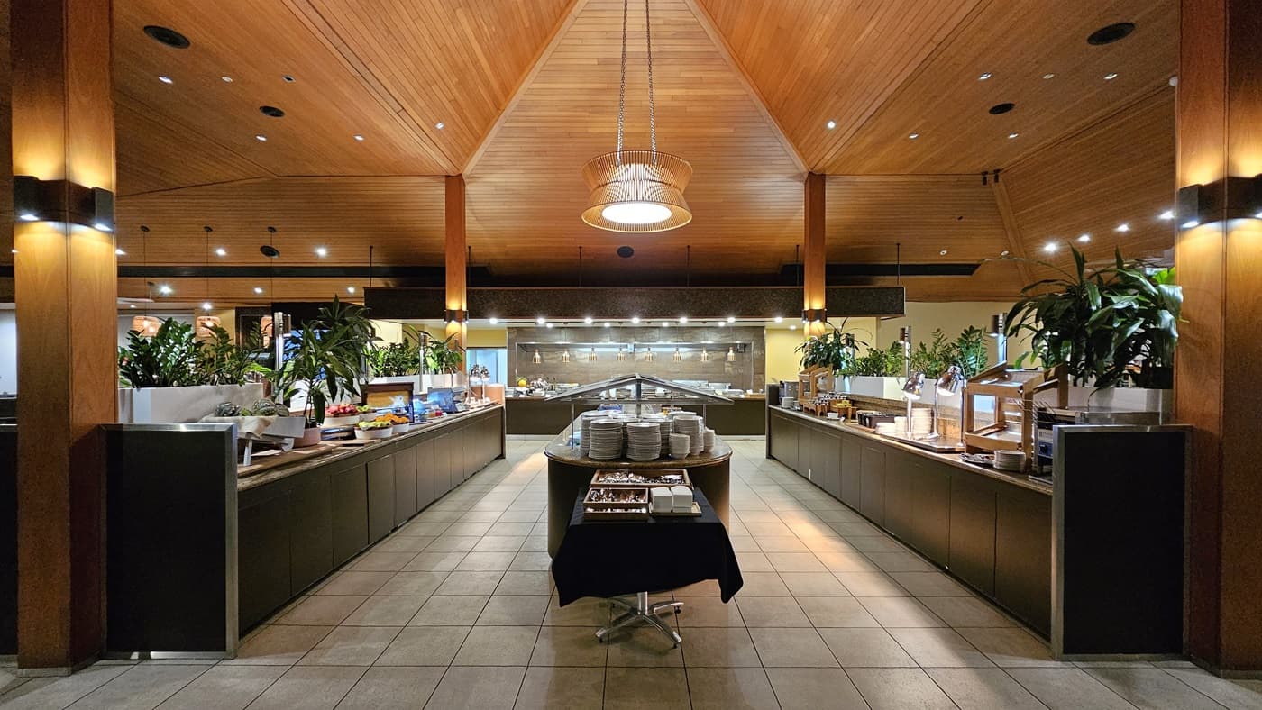 On the left, a long buffet counter with various food items and potted plants extends towards the back. To the right, a similar buffet counter mirrors the left side, also adorned with food items and plants. In the middle, a round table holds stacks of plates and cutlery. The background features a well-lit serving area with additional food options. The floor is tiled in light grey, and the ceiling is wooden with recessed lighting and a central pendant light.
