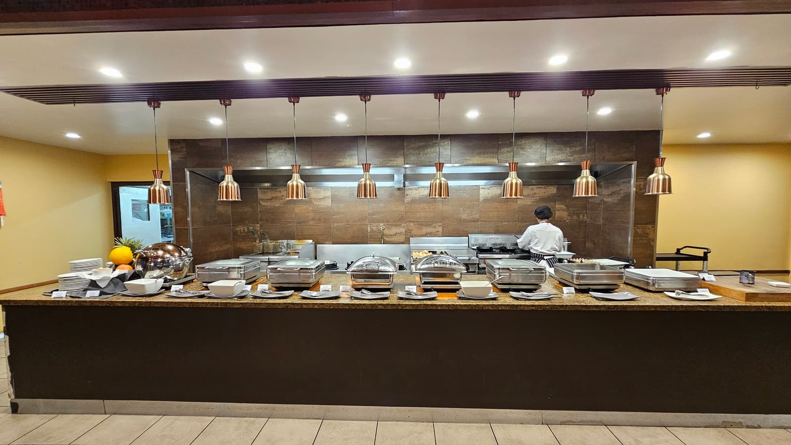 On the left, a stack of plates and bowls is arranged next to a fruit display on a granite counter. To the right, more serving dishes and utensils are placed on the same counter. In the middle, several chafing dishes with lids are lined up, with a chef working at a stove in the background. The floor is tiled in light grey, and the ceiling is white with recessed lighting and copper pendant lights hanging above the counter.