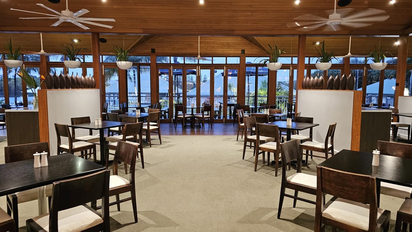 On the left, several dark wooden tables and chairs are arranged on a carpeted floor, with a white partition topped with decorative vases. To the right, a similar setup of tables and chairs is mirrored, also with a white partition and vases. In the background, large windows and glass doors open to an outdoor area, with hanging plants and ceiling fans above. The floor is a mix of carpet and dark wood, and the ceiling is wooden with recessed lighting and ceiling fans.