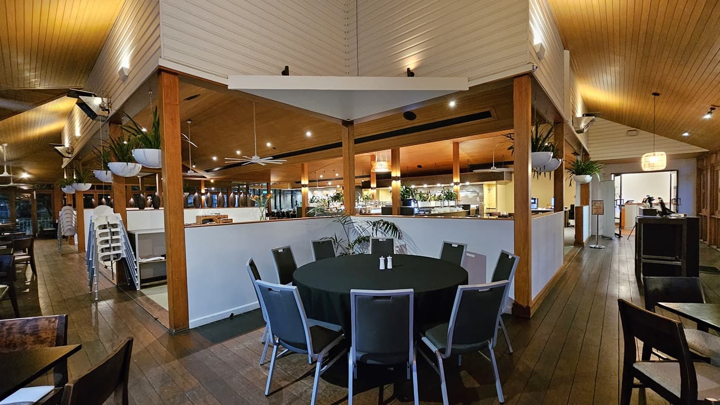 On the left, a row of dark wooden tables and chairs is arranged along a wooden floor, with stacked high chairs near a white partition. To the right, more tables and chairs are set up, with a reception area in the background. In the middle, a round table with a black tablecloth and grey chairs is placed on a carpeted section. The background features a buffet area with hanging plants and pendant lights. The ceiling is wooden with recessed lighting and ceiling fans.