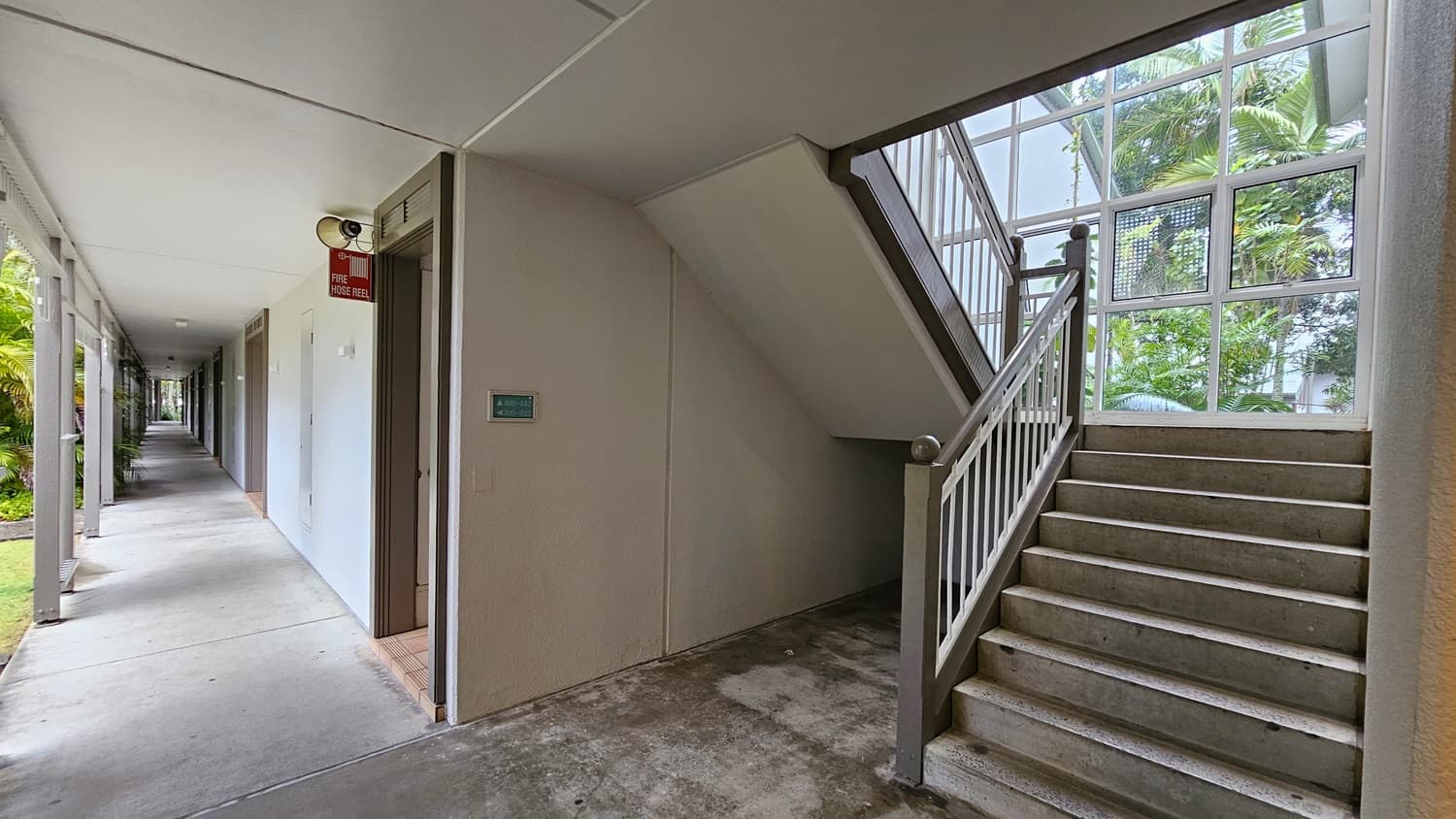 On the left, a long corridor with beige walls and grey doors extends into the distance, with a fire hose reel mounted on the wall. To the right, a staircase with grey steps and white railings ascends next to a large window with a view of green palm trees. In the middle, the corridor and staircase meet at a corner. The floor is concrete, and the ceiling is flat and white with recessed lighting.