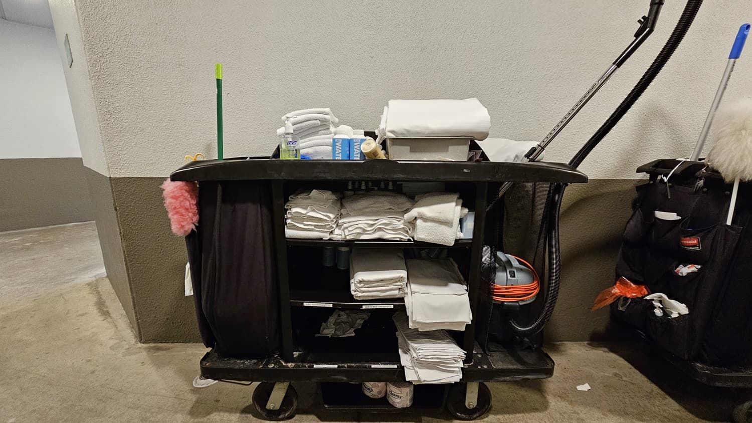 On the left, a beige and grey wall with a green broom leaning against it. To the right, a black cleaning cart with shelves filled with folded white towels, cleaning supplies, and a vacuum cleaner with an orange cord. In the middle, more folded towels and a box of tissues are on top of the cart.
