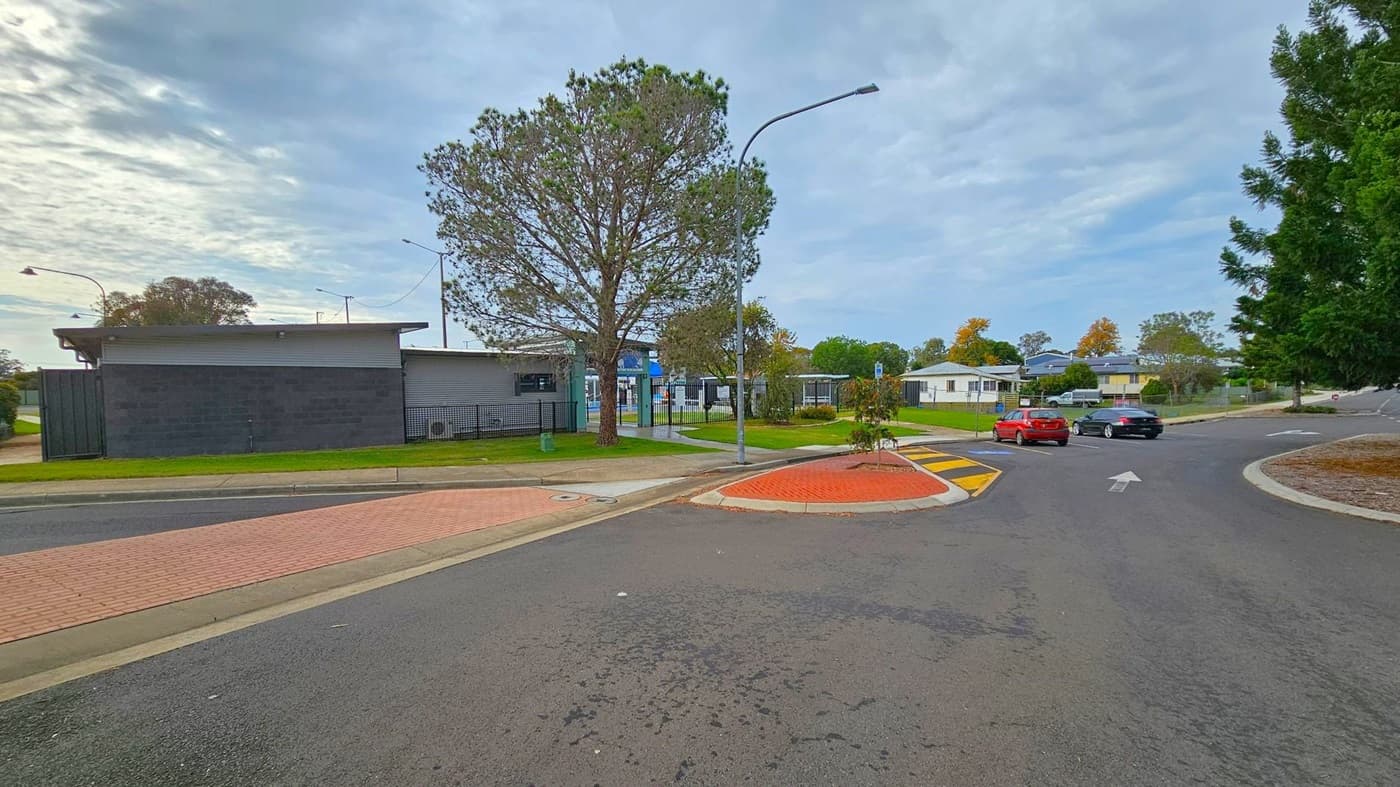 A street scene with a black building on the left, featuring a flat roof and surrounded by a grassy area. A large tree stands nearby, next to a metal fence. In the centre, a road curves to the right, with a red car and a black car driving away. On the right, there are more trees and a few residential buildings in the background. The road has a yellow and black speed bump and a red brick pedestrian crossing. Street lamps are visible along the road.