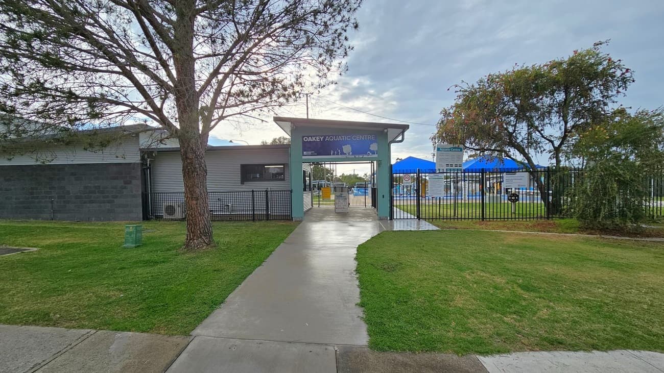 A pathway leads to the entrance of the Oakey Aquatic Centre, flanked by a large tree on the left and a smaller tree on the right. The building on the left has a grey exterior with a window. A black metal fence surrounds the area, with signs visible near the entrance. Blue shade structures can be seen in the background, indicating a pool area. The ground is covered with grass and concrete paths.