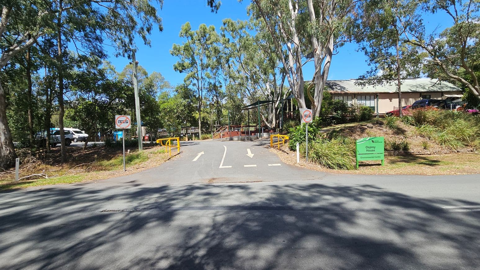 A paved road leads to a building partially visible on the right, surrounded by trees and greenery. Yellow barriers and directional arrows are on the road. Two signs indicate no U-turns. A green sign on the right reads "Osprey House Environmental Centre." On the left, a parked white vehicle is visible among the trees. The area is shaded by tall trees, and the sky is clear and blue.