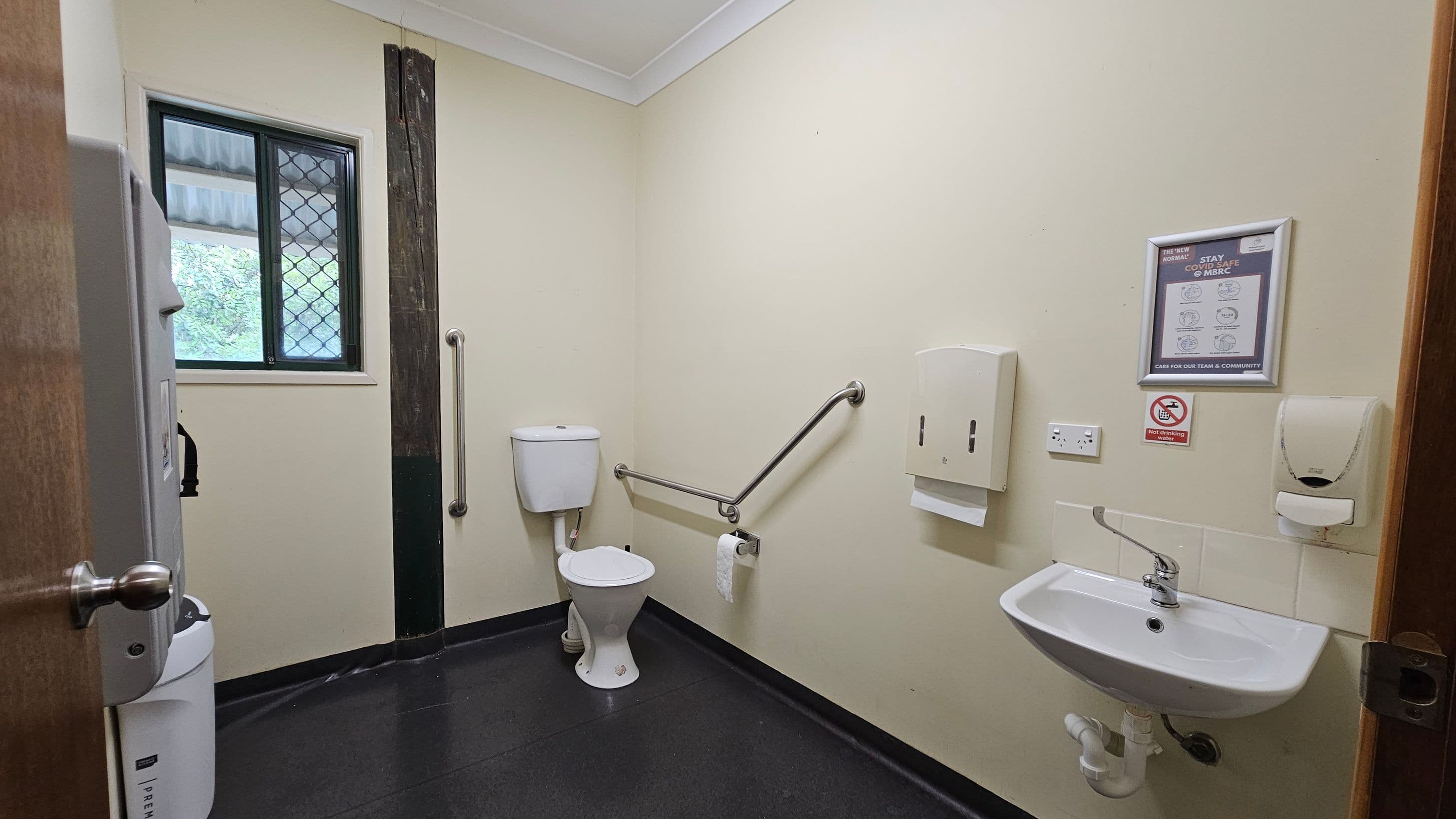 A public restroom with a white toilet and grab bars on the left. A small window with a security grille is above the toilet. On the right, a white sink is mounted on the wall with a soap dispenser and paper towel holder nearby. A framed sign and a no-smoking notice are above the sink. The floor is dark and the walls are light-coloured. The ceiling is white with a simple design.