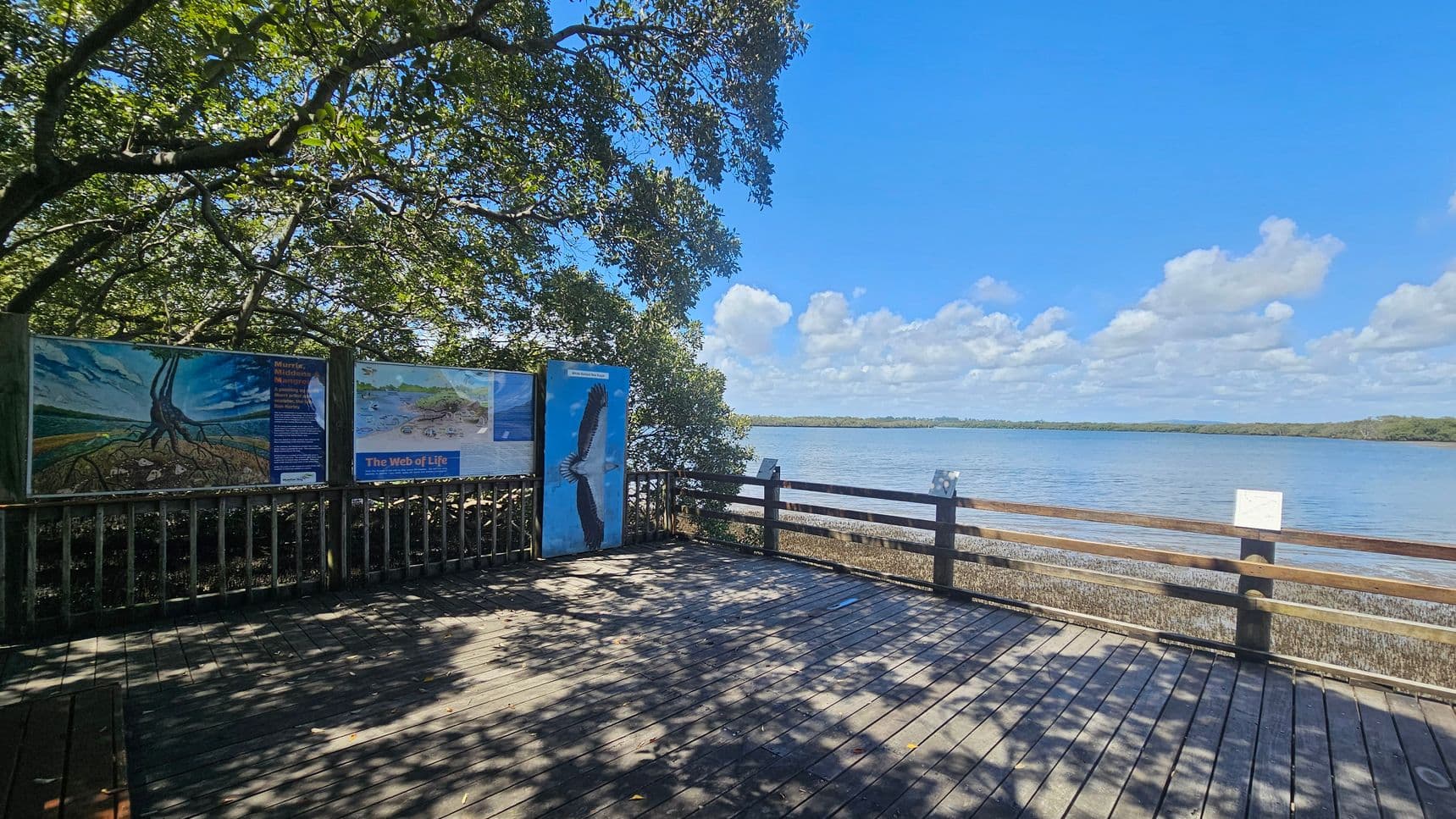 A wooden deck overlooks a body of water with a railing on the right. On the left, there are informational signs with images, including one of a bird. Trees with dense foliage provide shade, casting shadows on the deck. The background features a calm water surface extending to a distant tree-lined shore under a blue sky with scattered clouds.
