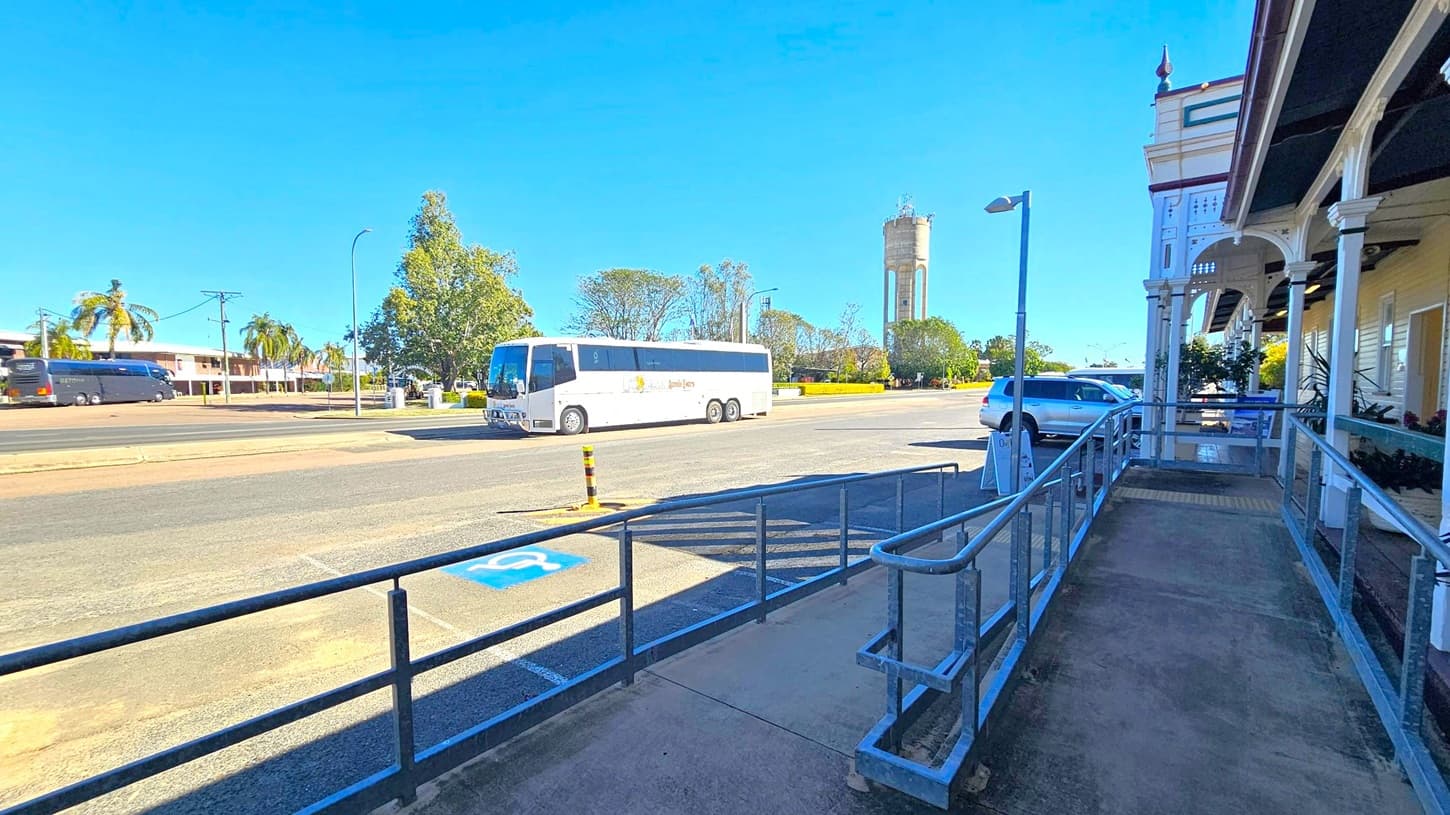 A street scene with a white tour bus parked on the left side of the road. In the background, there are palm trees, a large tree, and a tall water tower. On the right, a building with a covered walkway and decorative columns is visible. A silver car is parked nearby, and a ramp with metal railings leads up to the building. The pavement has a marked disabled parking space. The sky is clear and blue.