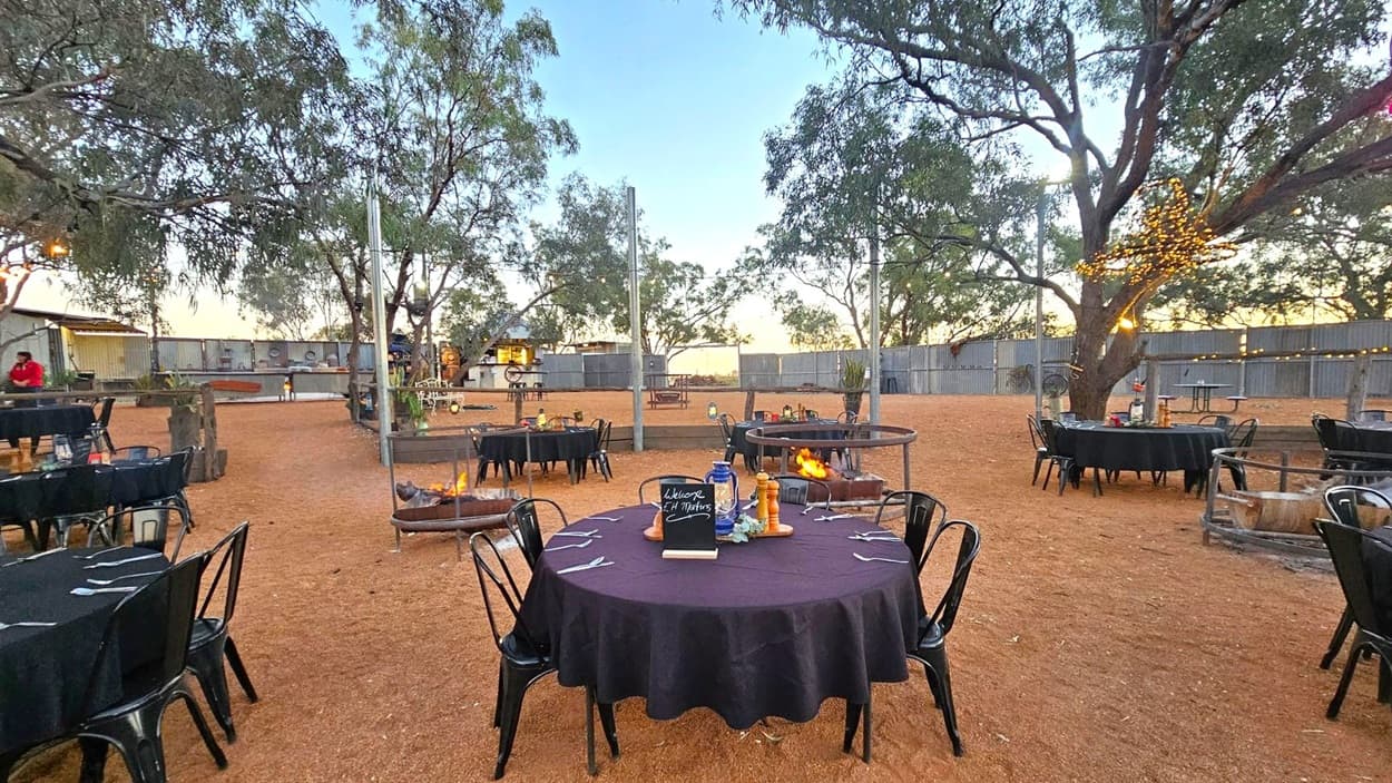 Outdoor dining area with round tables covered in black tablecloths, surrounded by black chairs. In the centre, a table displays a small chalkboard and condiments. Fire pits are scattered around, with one in the foreground. Trees with string lights are visible, and a wooden fence encloses the space. The ground is covered in reddish-brown dirt, and there are additional tables and a structure in the background.
