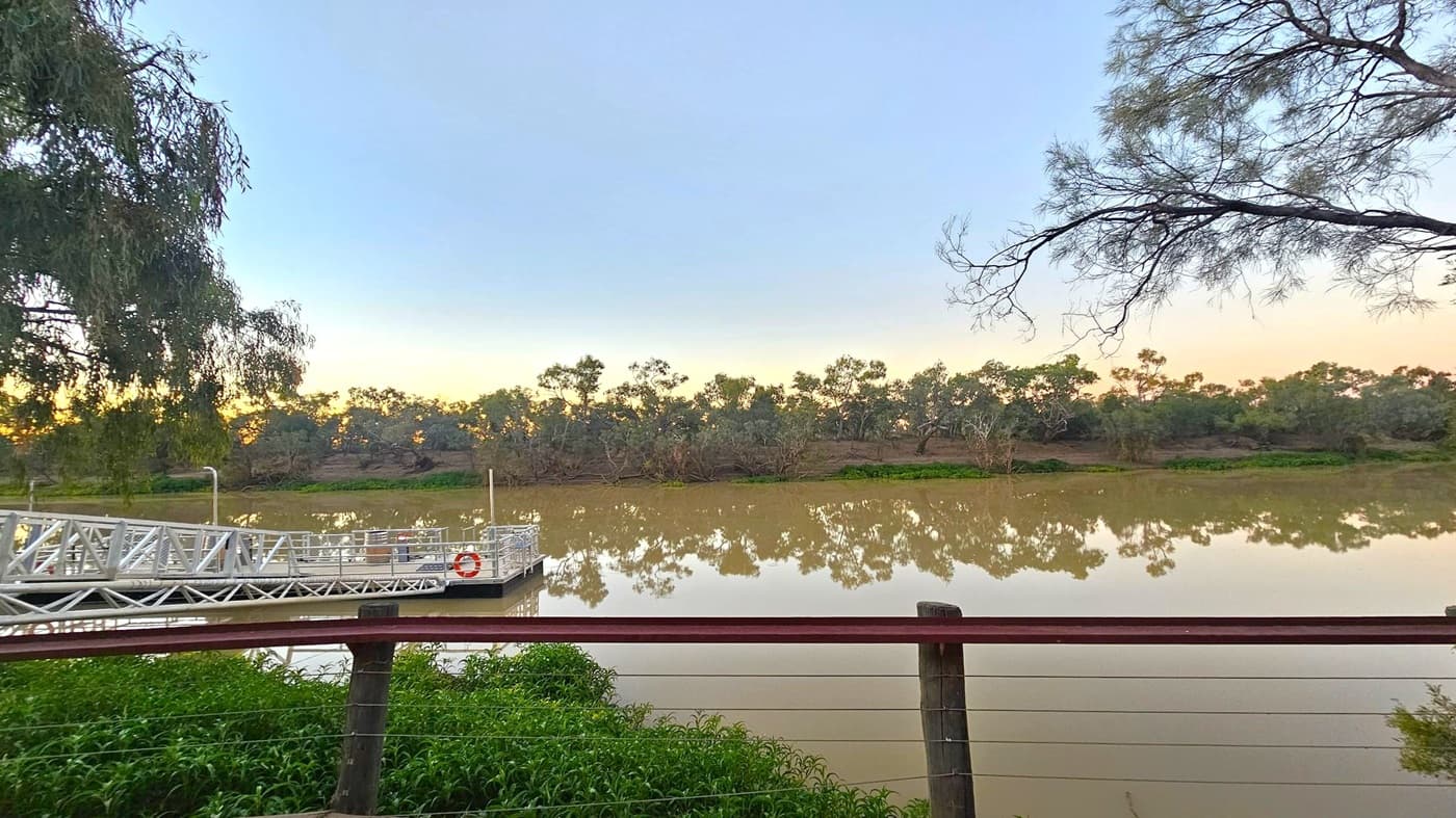 A calm river with a reflection of trees along the opposite bank. In the foreground, a metal railing and a small dock with a lifebuoy are visible. The dock extends into the water from the left. Trees with sparse branches frame the scene on both sides. The water is a muddy brown, and the sky has a gradient of light blue to pale orange near the horizon. Green foliage is visible near the railing.