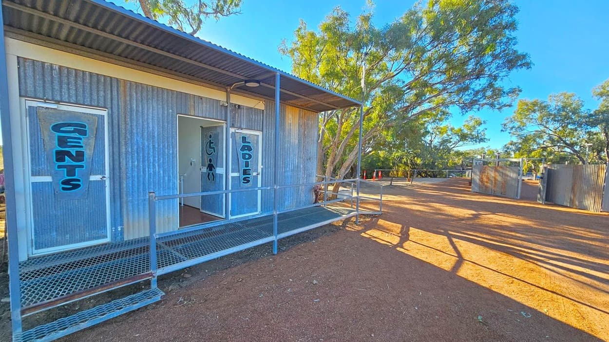 A corrugated metal building with two doors labelled "Gents" and "Ladies" on the left. The building has a metal ramp leading to the entrances. The ground is covered with reddish-brown dirt. In the background, there are trees and additional metal structures. The area is well-lit, suggesting daytime. Shadows from the trees and structures stretch across the ground.