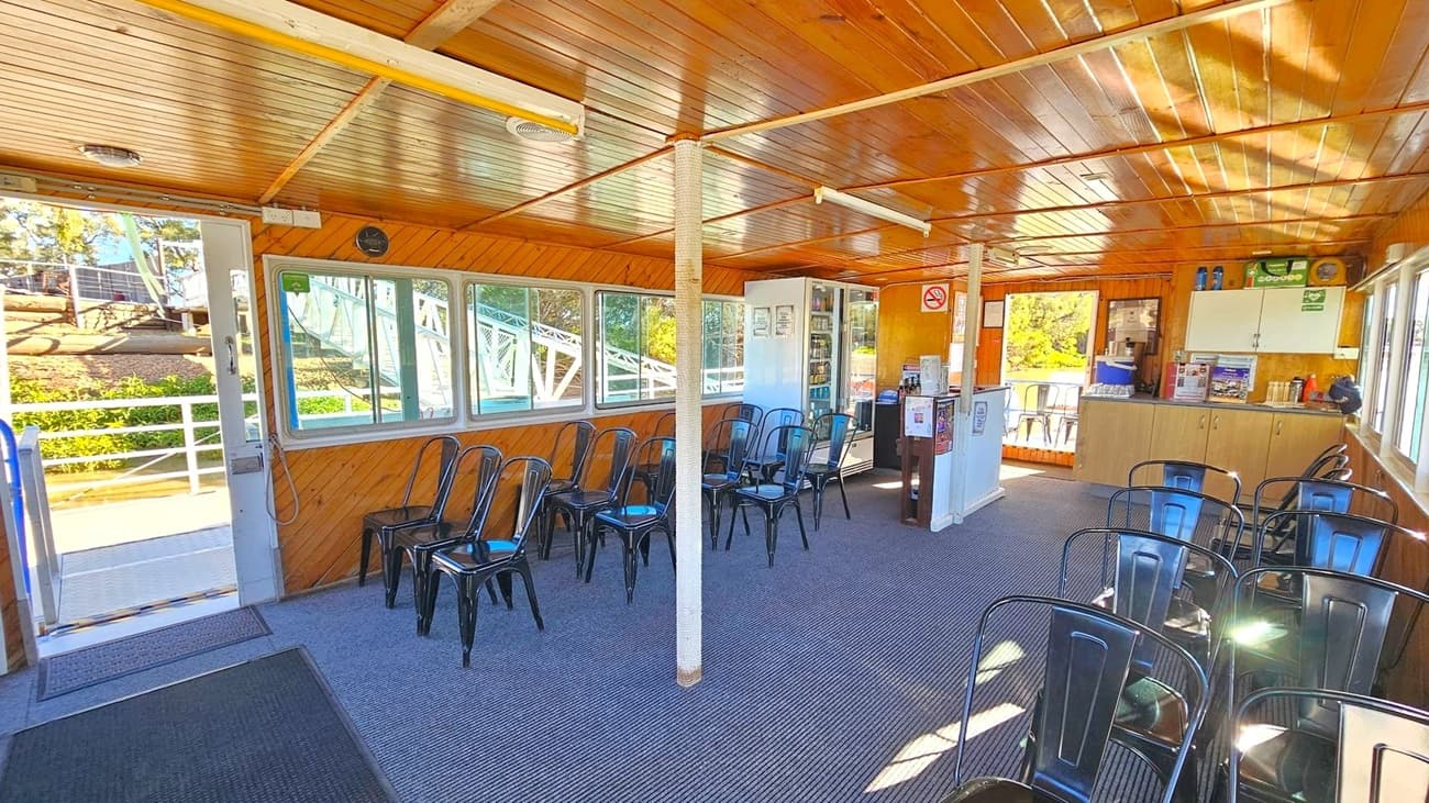 Interior of a wooden cabin with a slatted ceiling and carpeted floor. On the left, a door opens to an outdoor area with a view of trees and a railing. Several black metal chairs are arranged along the walls and in the centre. On the right, a counter with cabinets above and a small fridge below holds various items. Windows line the walls, allowing natural light to illuminate the space. A white column stands in the middle of the room.