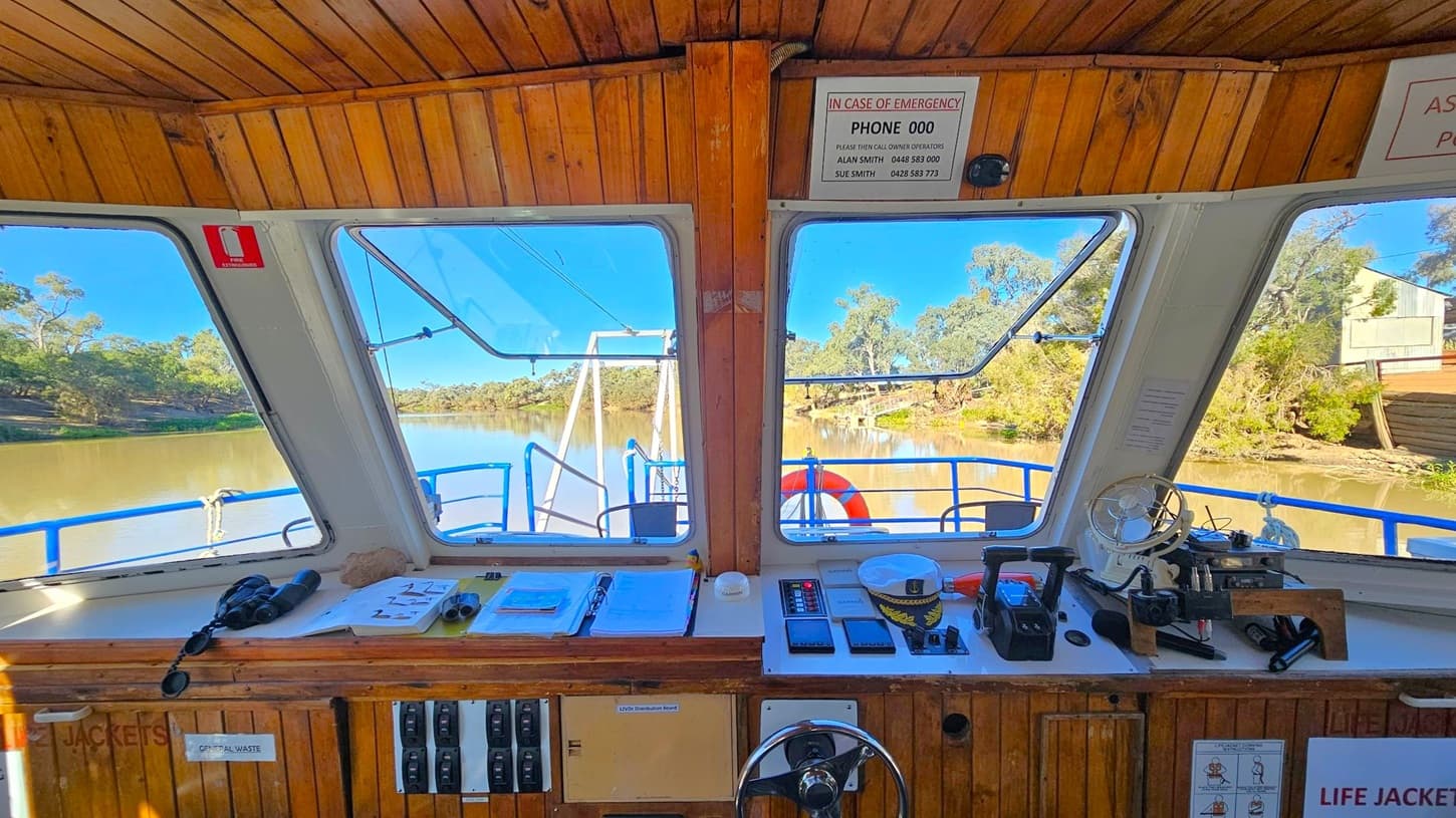 The image shows the interior of a boat's control cabin. The foreground features a wooden dashboard with various controls, switches, and navigation equipment. Papers, a pair of binoculars, and a small fan are also visible. Through the large windows, a river with trees lining the banks can be seen. A life ring is attached to the boat's railing outside. The ceiling is wooden, and emergency contact information is displayed above the windows.