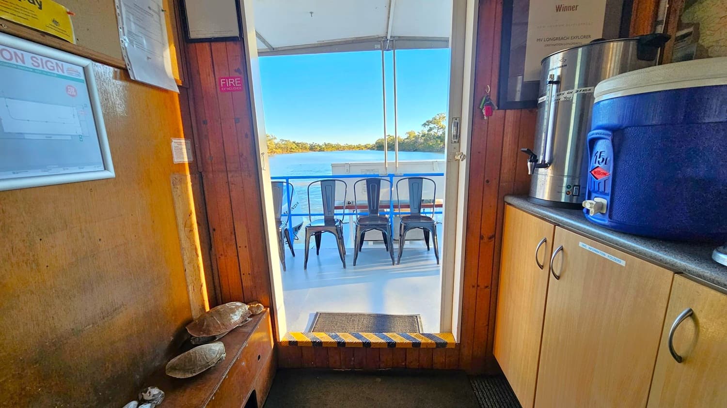 Wooden interior with a notice board on the left wall displaying papers and a fire extinguisher sign. A bench below holds several shells. On the right, a countertop with a large metal urn and a blue water cooler sits atop light-coloured cabinets. Through the open door, a view of a deck with metal chairs facing a body of water and trees in the distance. The floor is carpeted inside and transitions to a smooth surface outside.