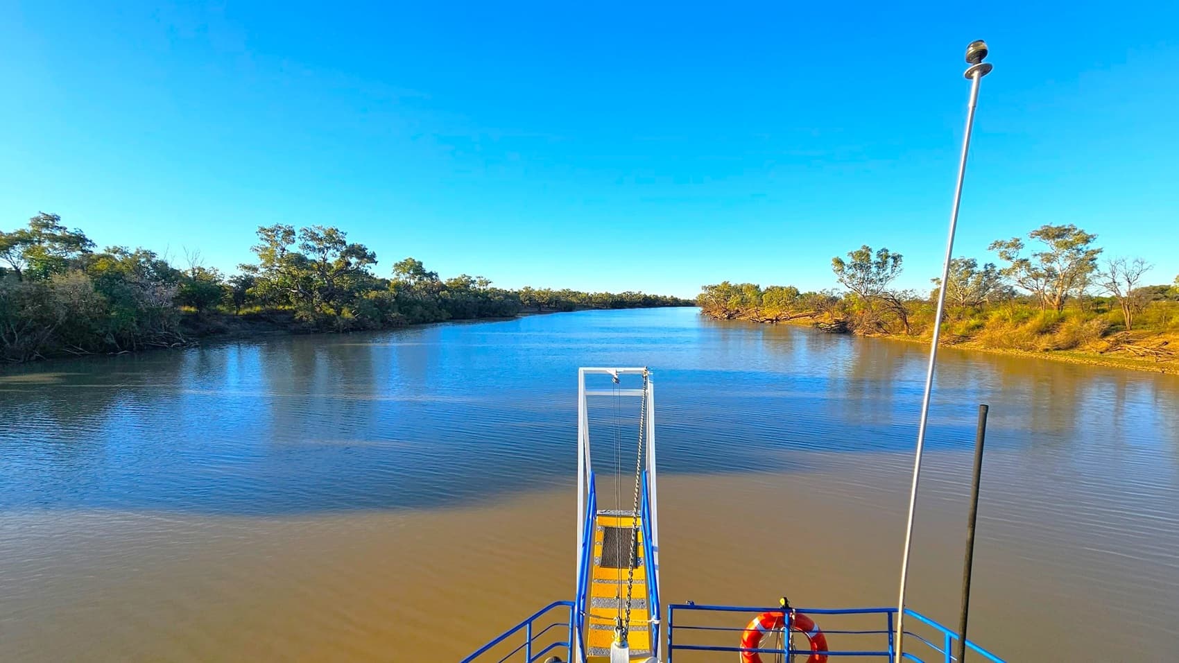 A wide river stretches into the distance, flanked by dense green trees on both sides. In the foreground, a blue and yellow metal structure with a life buoy is visible, possibly part of a boat or dock. A tall metal pole with a device on top extends upwards. The water is calm, reflecting the clear blue sky above. The riverbanks are lined with vegetation, and the scene is tranquil and expansive.