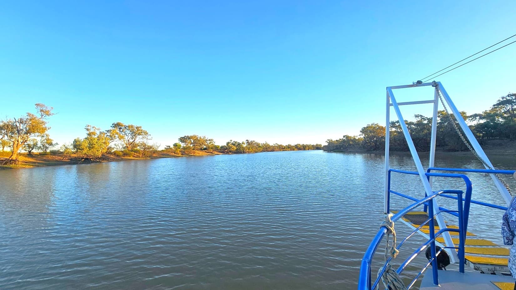 A calm river stretches into the distance, flanked by trees and low vegetation on both sides. The water reflects the surrounding landscape. On the right, part of a boat is visible with blue railings and a white metal structure. A rope is tied to the railing, and a patterned fabric is draped over it. The scene is serene, with the natural environment dominating the view.