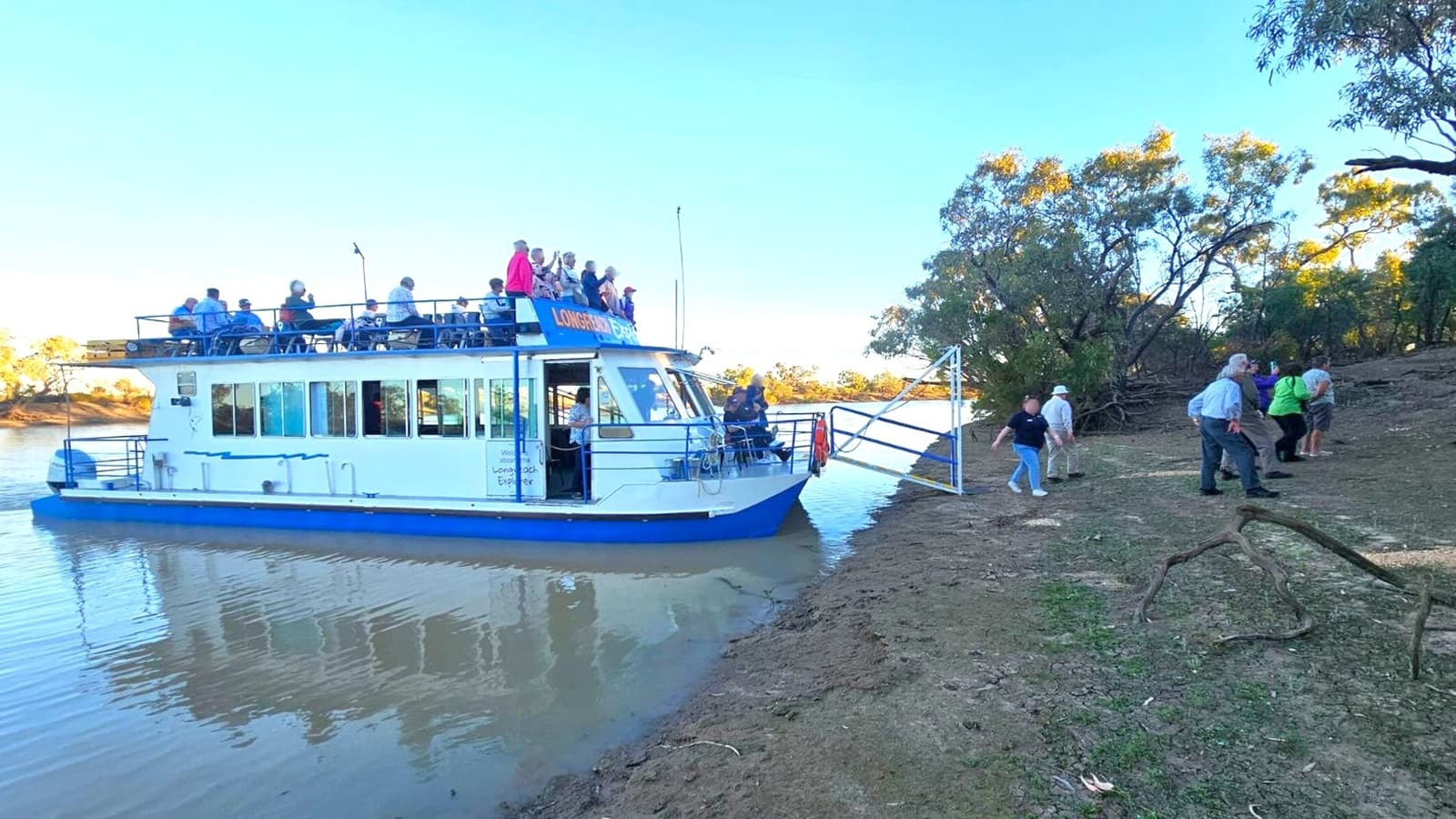 A boat with a blue and white exterior is docked on a riverbank. People are seated on the upper deck, while others are disembarking onto the muddy shore. The riverbank is uneven with patches of green vegetation and scattered branches. Trees with sparse foliage are visible in the background, and the water is calm with gentle ripples. The sky is clear and blue.