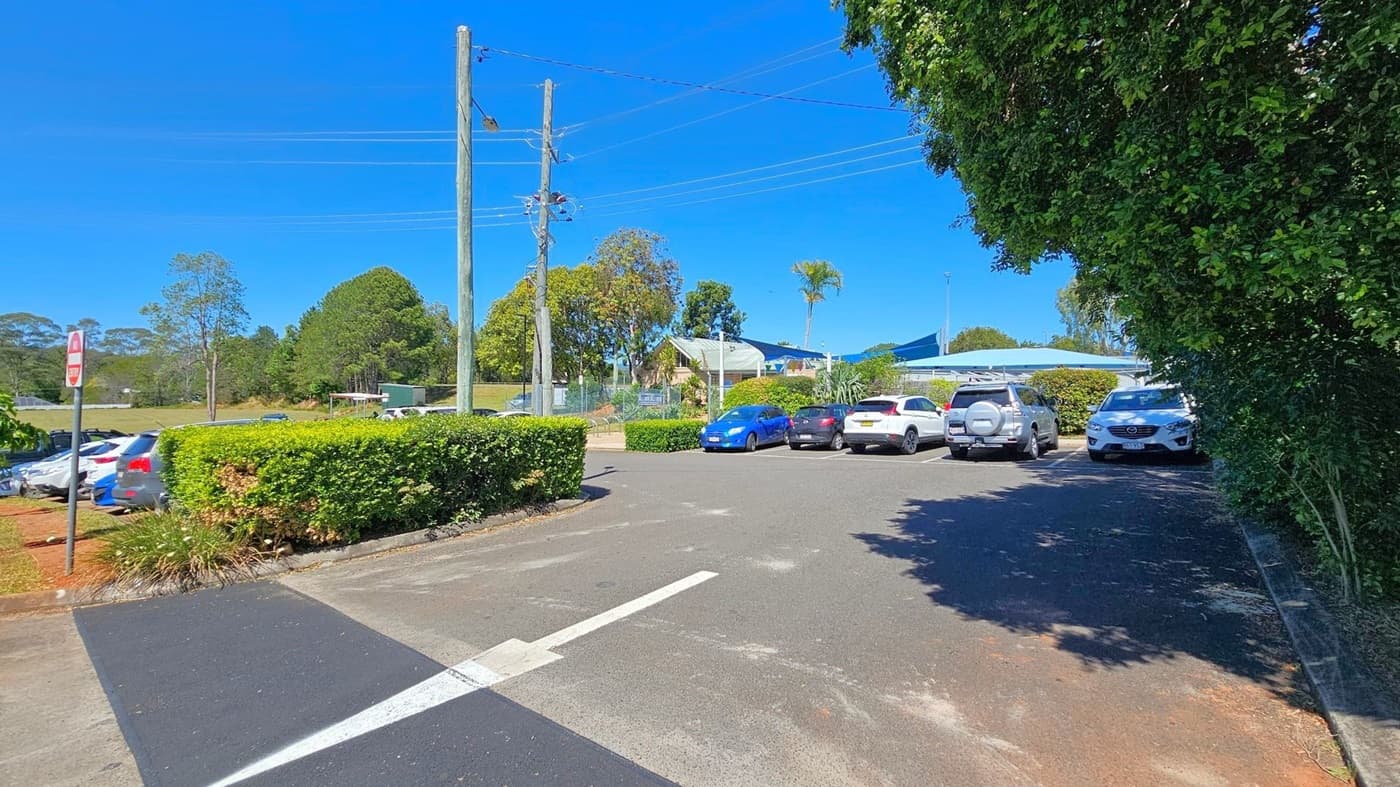 A car park with several vehicles, including sedans and SUVs, is on the left side. Large trees provide shade over some of the cars. In the middle, a paved road with a white arrow pointing forward is visible. On the right, more parked cars are partially obscured by a neatly trimmed hedge. Utility poles with power lines run along the right side, and a building with a green roof is in the background. The ground is paved, and the sky is clear.