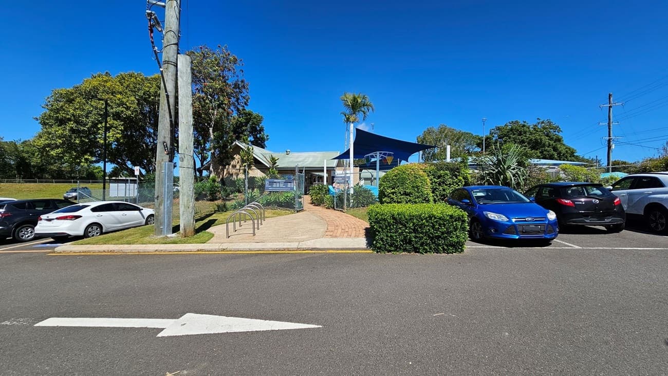 A paved pathway leads to a building with a green roof and a blue shade structure. The entrance is flanked by a chain-link fence and greenery. On the left, there is a row of parked cars, including a white car. On the right, there are more parked cars, including a blue car and a black car. A large utility pole stands near the centre. The background features trees and additional greenery. The foreground shows an asphalt road with a white arrow pointing forward.