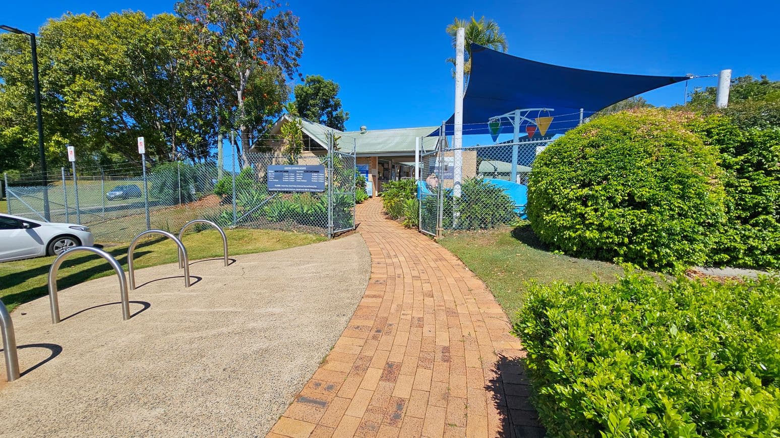 A brick pathway leads to a gated entrance with a sign displaying information. On the left, there is a bike rack and a white car parked near a chain-link fence. Behind the fence, there are large trees with green foliage. On the right, there are well-maintained bushes and a shaded area with a blue canopy. The background features a building with a green roof and more greenery. The sky is clear and blue.