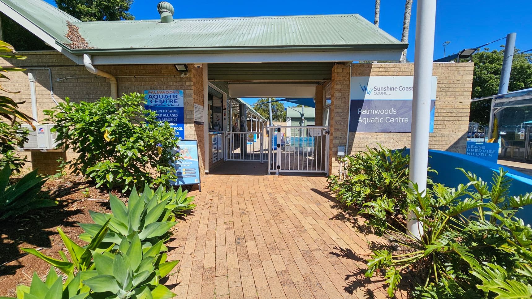 Entrance to Palmwoods Aquatic Centre with a brick pathway leading to a gated entry. On the left, there are green plants and a sign listing services like swimming lessons and aqua aerobics. The building has light-coloured brick walls and a green metal roof. On the right, more green plants and a sign with the centre's name. A white pole is also visible on the right side. In the background, there are more buildings and a shaded area.