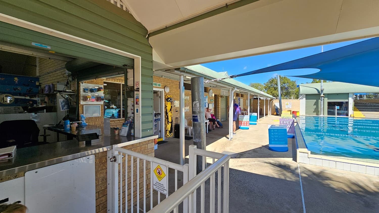 A poolside area with a green-roofed building on the left, featuring a service window with various items and a counter. In the middle, there is a walkway with a first aid station and a yellow spine board mounted on the wall. People are seated on benches along the walkway. On the right, there is a swimming pool with blue shade sails overhead. The floor is concrete, and the ceiling is a combination of the building's roof and the shade sails.
