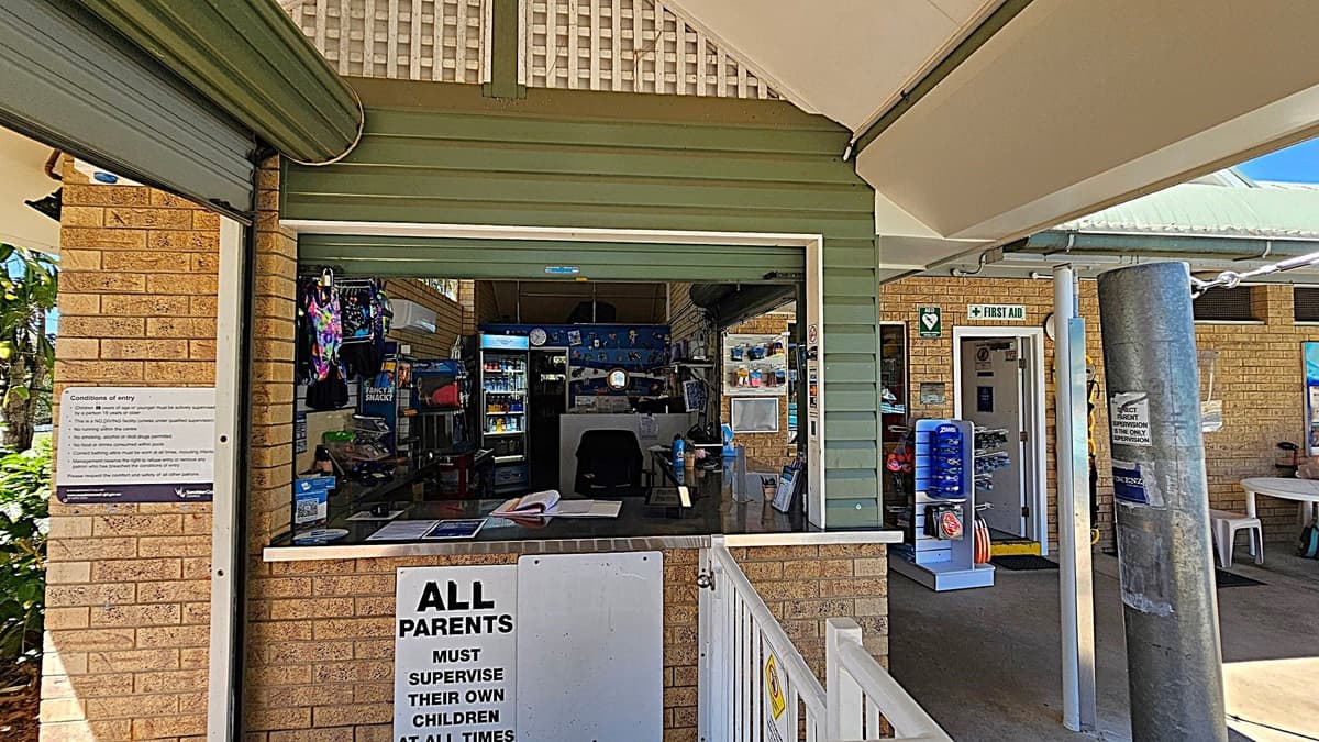 A kiosk with a green and brick exterior. On the left, a sign lists the conditions of entry. In the middle, the kiosk window displays various items for sale, including snacks and swim gear. Behind the counter, a refrigerator with drinks is visible. On the right, a first aid sign is above a door leading to a room with more items. A large grey post stands in front of the kiosk, and a white fence with a sign about parental supervision is in the foreground.
