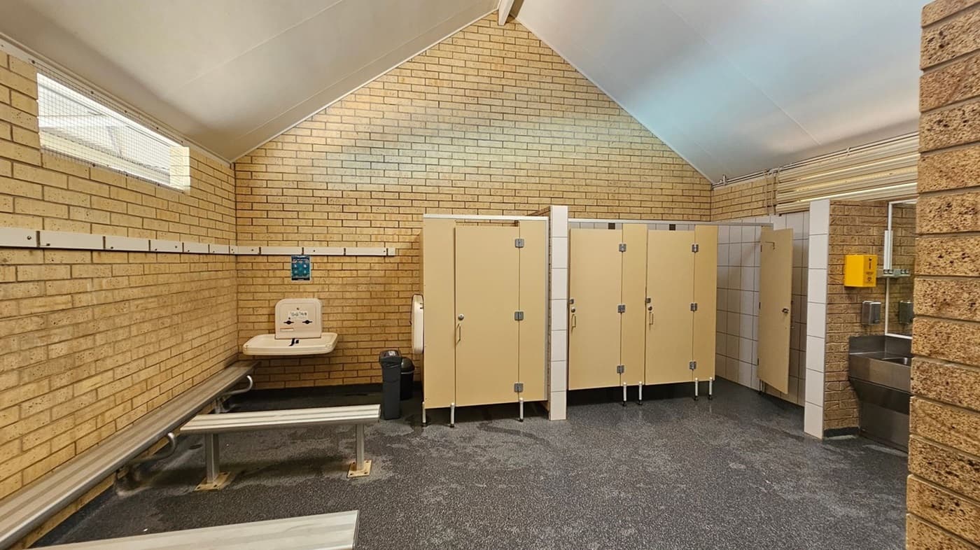 A changing room with beige brick walls and a high, slanted white ceiling. On the left, there are metal benches and a baby changing station mounted on the wall. In the middle, there are beige-coloured toilet cubicles with closed doors. On the right, there is a stainless steel sink with a yellow soap dispenser above it. The floor is covered with a speckled blue and green surface. High windows with wire mesh are present on the left wall.