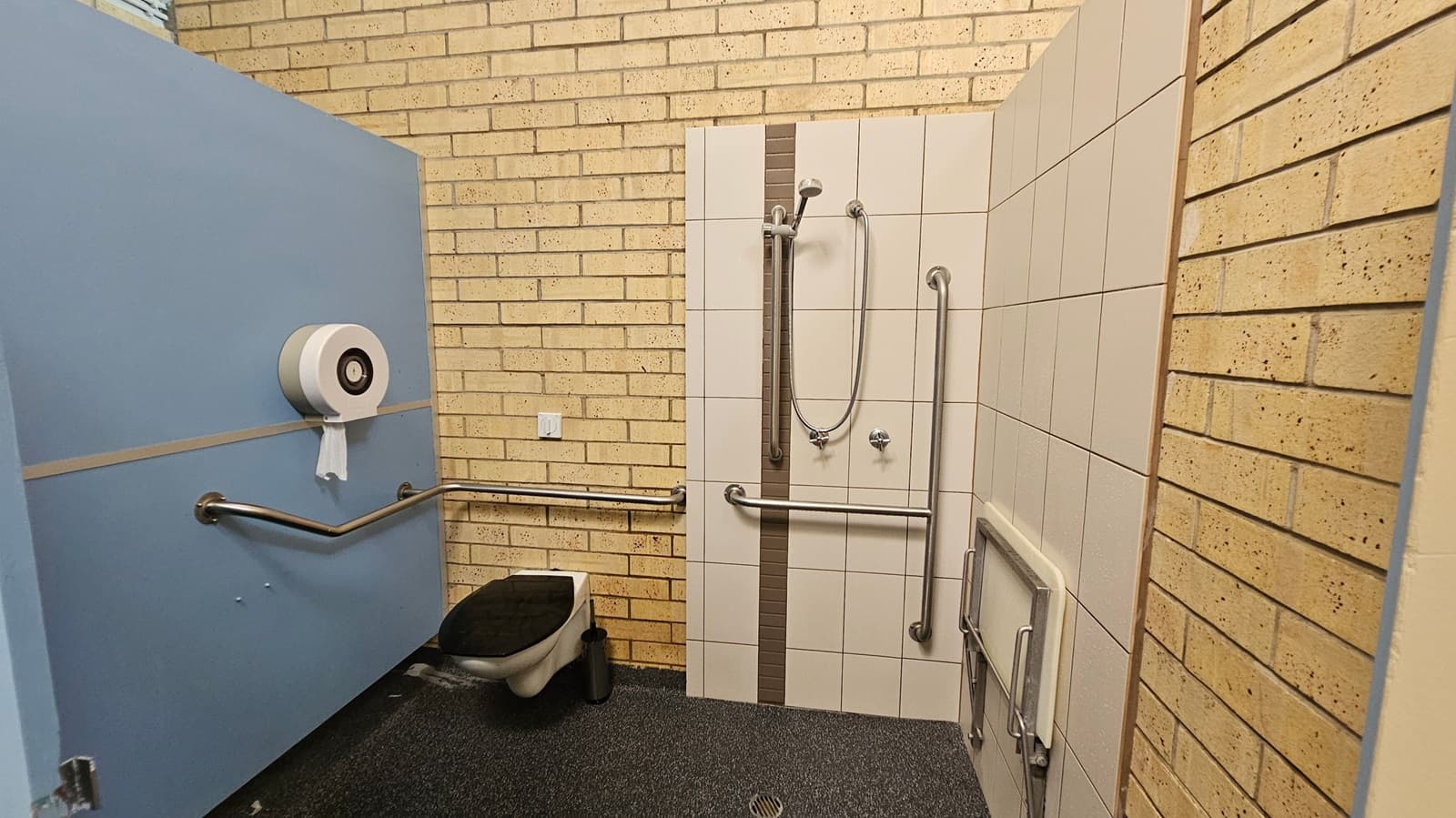 Accessible bathroom with light yellow brick walls and a blue door on the left. The door has a large toilet paper dispenser and a horizontal grab bar. The toilet is positioned against the left wall with a black seat. The shower area is in the middle, featuring white tiles with a vertical grey stripe, a handheld showerhead, and multiple grab bars. The floor is covered in a speckled blue and green non-slip material. A foldable shower seat is attached to the right wall.