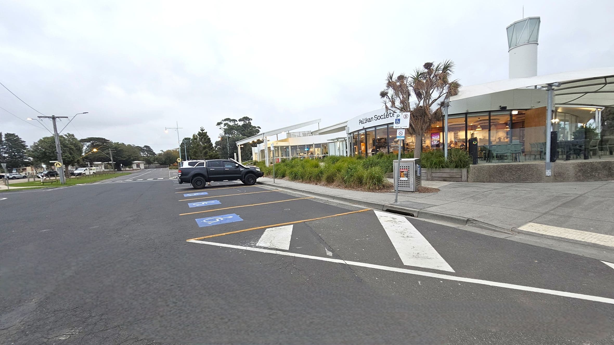 A car park with designated disabled parking spaces marked in blue is on the left. A black vehicle is parked in one of the spaces. On the right, a modern building with large glass windows and a sign reading "Pelikan Societ" is visible. The building is surrounded by landscaped greenery, including small trees and shrubs. A rubbish bin is placed near the entrance. The pavement is grey, and a tall structure with a glass top is in the background.