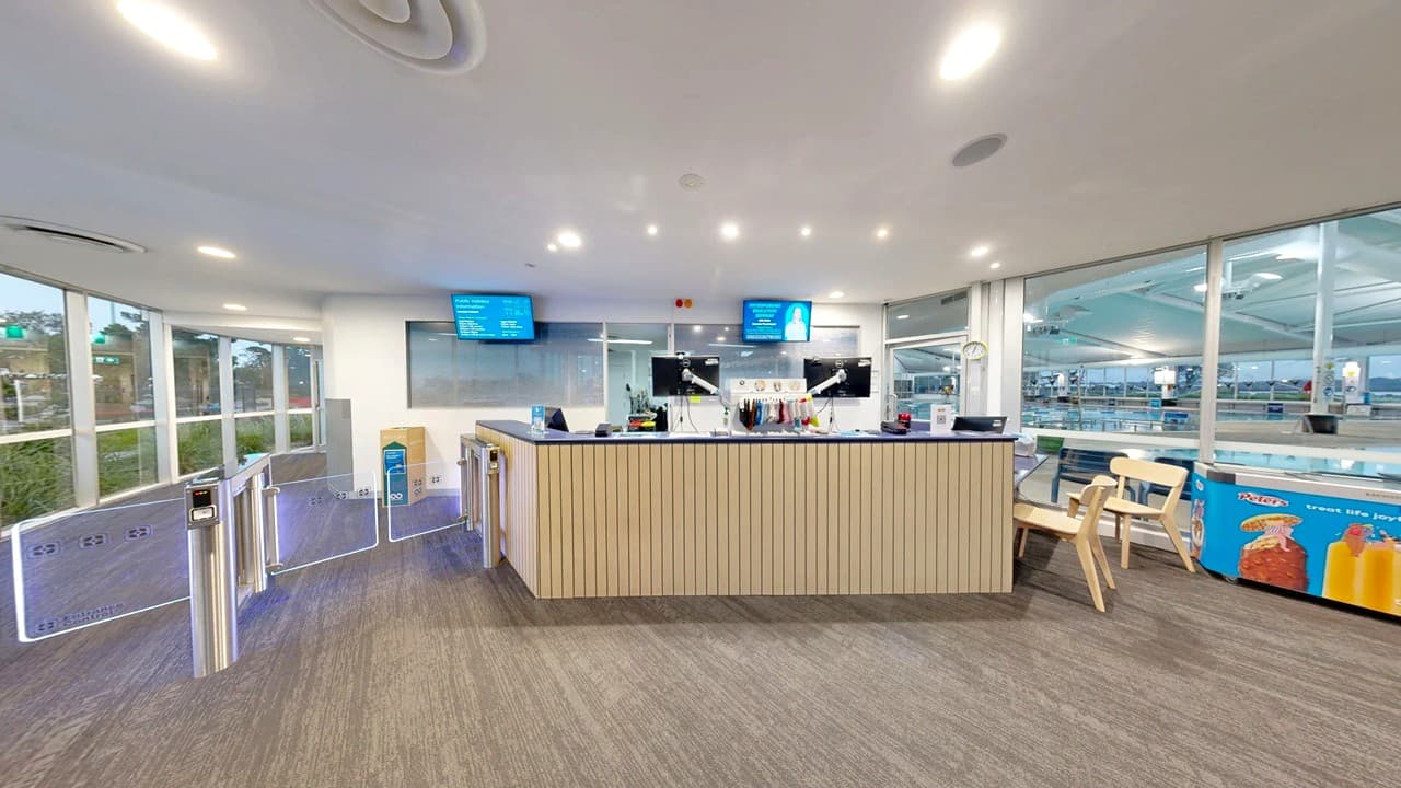 A modern reception area with a wooden counter in the centre, equipped with computers and microphones. Two digital screens are mounted on the wall behind the counter. On the right, there are wooden chairs and a display with snacks and drinks. The left side features a series of turnstiles leading to a glass wall with a view of greenery outside. The floor is carpeted, and the ceiling has recessed lighting.
