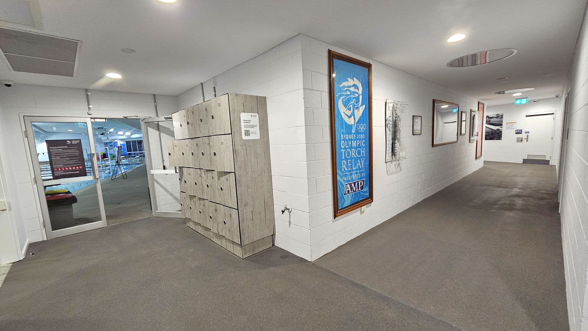 A hallway with white brick walls and a brown and grey carpet. On the left, there are wooden lockers and a glass door leading to a swimming pool area. The right wall features framed posters, including one for the Sydney 2000 Olympic Torch Relay. The ceiling is white with recessed lighting, and there is a circular vent. At the end of the hallway, there is a door with a sign and an emergency exit light above it.