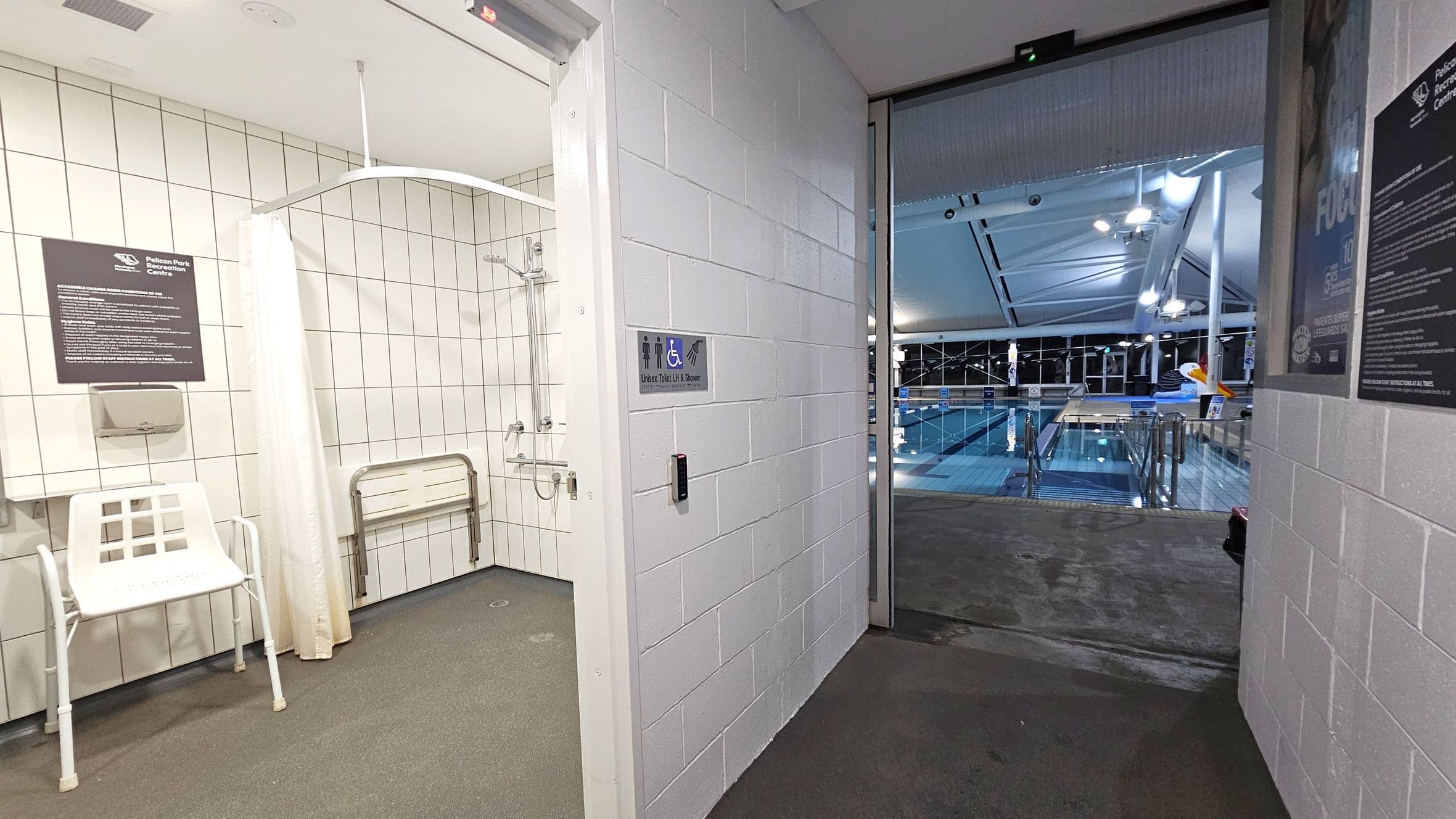 On the left, there is a tiled room with a shower, a white chair, and a folding bench. A sign and a paper towel dispenser are mounted on the wall. In the centre, a door leads to a pool area. The wall beside the door has a sign indicating a unisex toilet and shower. On the right, the pool area is visible with a large swimming pool, metal railings, and a high ceiling with lights. The floor is a mix of tiles and concrete.