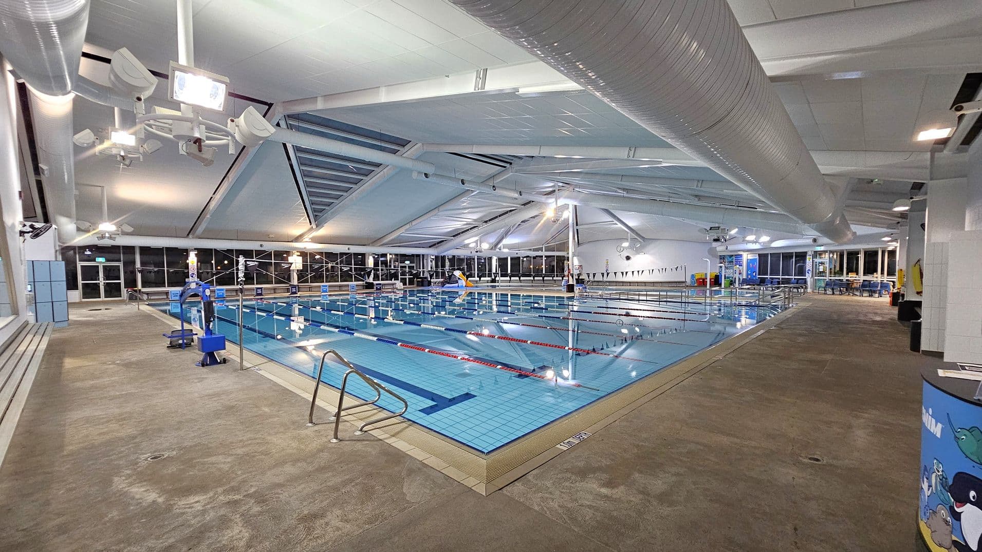 An indoor swimming pool with clear blue water is in the centre, divided into lanes by red and blue ropes. On the left, there are blue lockers and a bench along the wall. The right side features a wide walkway with a colourful cylindrical bin. The ceiling is high with large white ducts and bright lighting. The floor is a textured, non-slip surface. Large windows line the background, allowing natural light to enter.