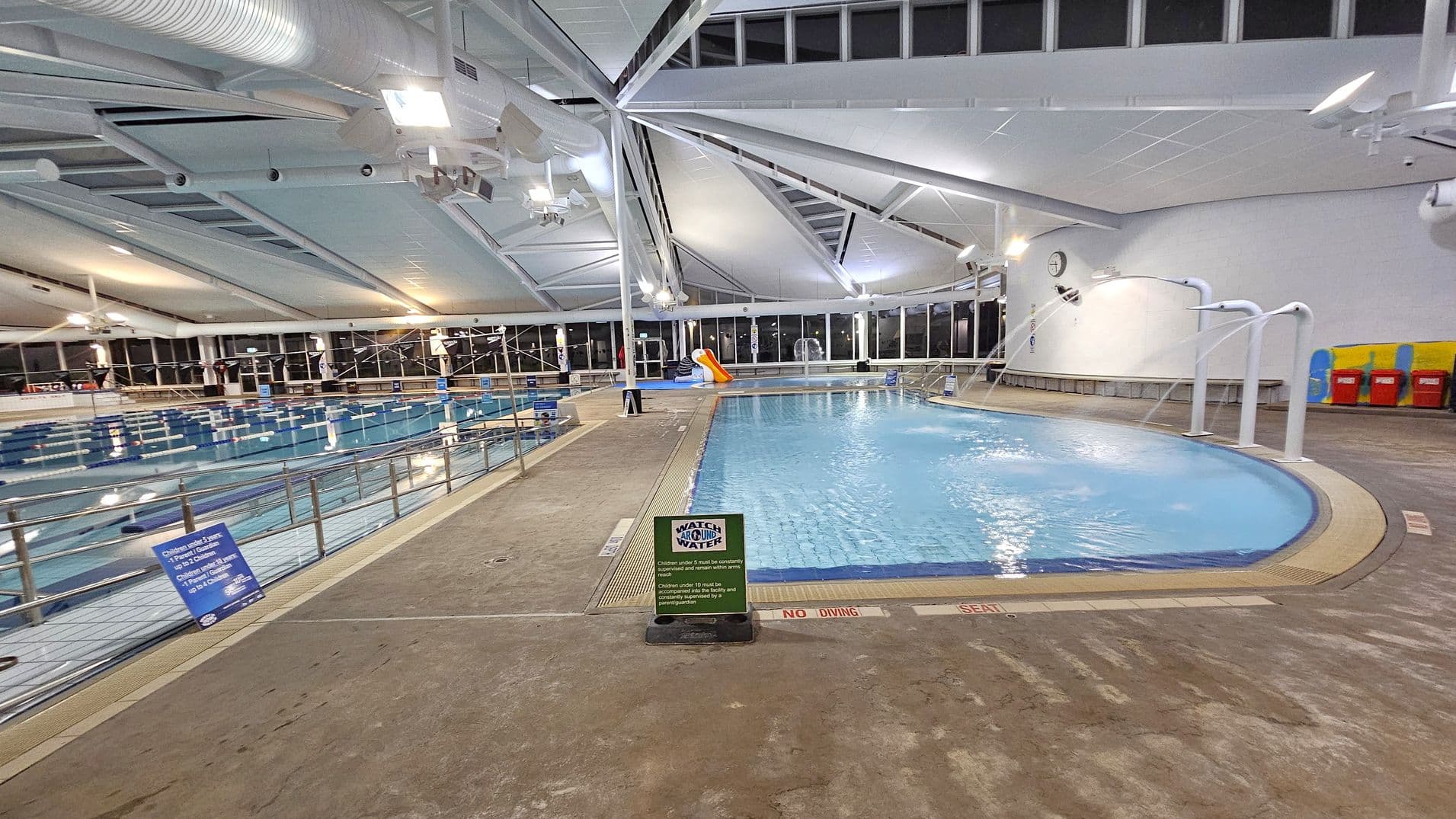 Indoor swimming pool area with two pools. On the left, a large lap pool with lane dividers and a railing along the edge. On the right, a smaller leisure pool with water features and a slide. The floor is textured concrete with safety signs. The ceiling is high with exposed beams and ductwork, and large windows line the walls, allowing natural light. A clock is mounted on the wall above benches and colourful lockers.