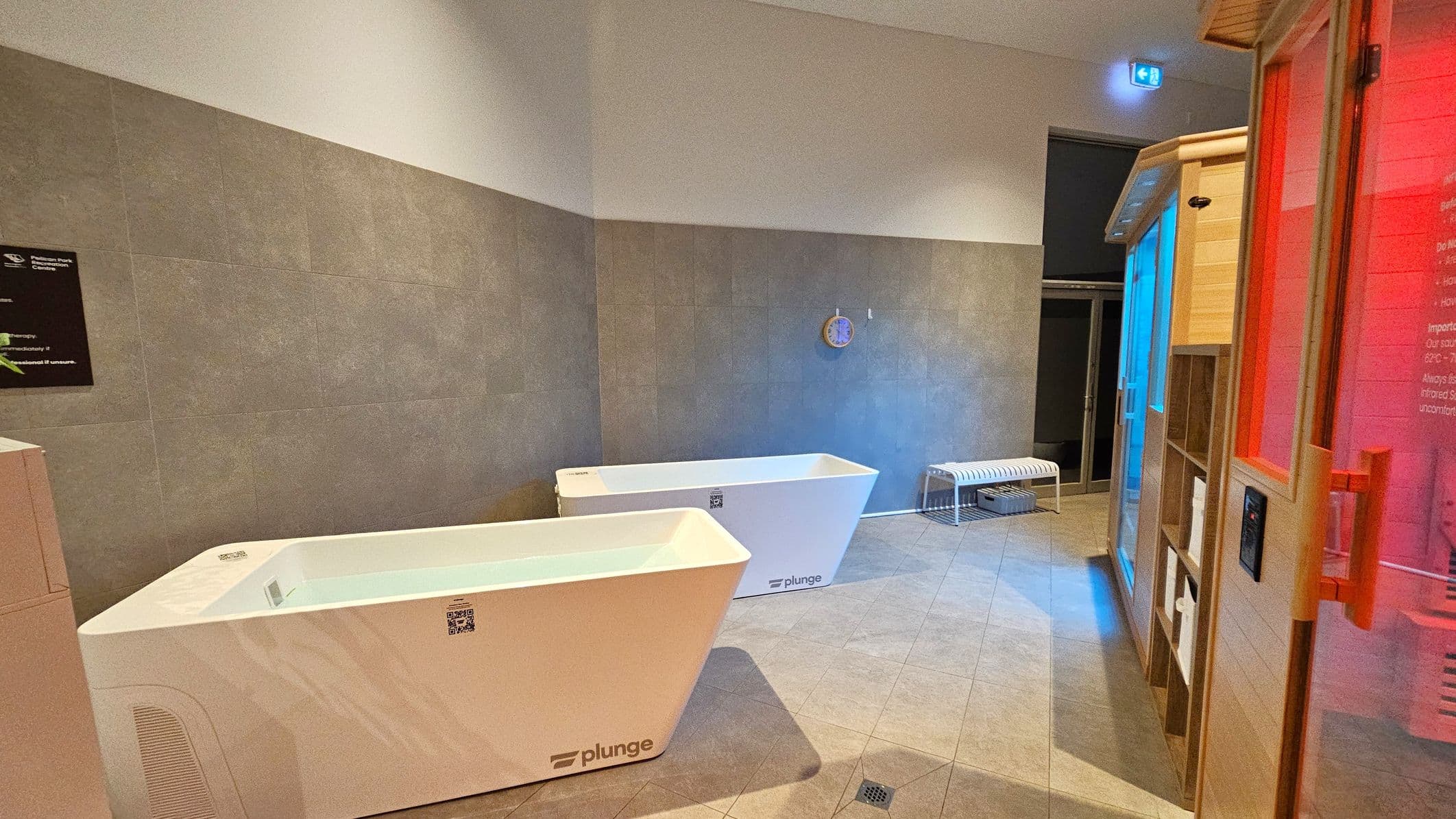 On the left, there are two white plunge tubs with a modern design, placed against a grey-tiled wall. In the middle, a small clock is mounted on the wall above a white bench. On the right, a wooden sauna with a glass door emits a red glow, and a shelf unit is visible beside it. The floor is tiled in a light colour, and the ceiling is plain with a blue exit sign above the door in the background.