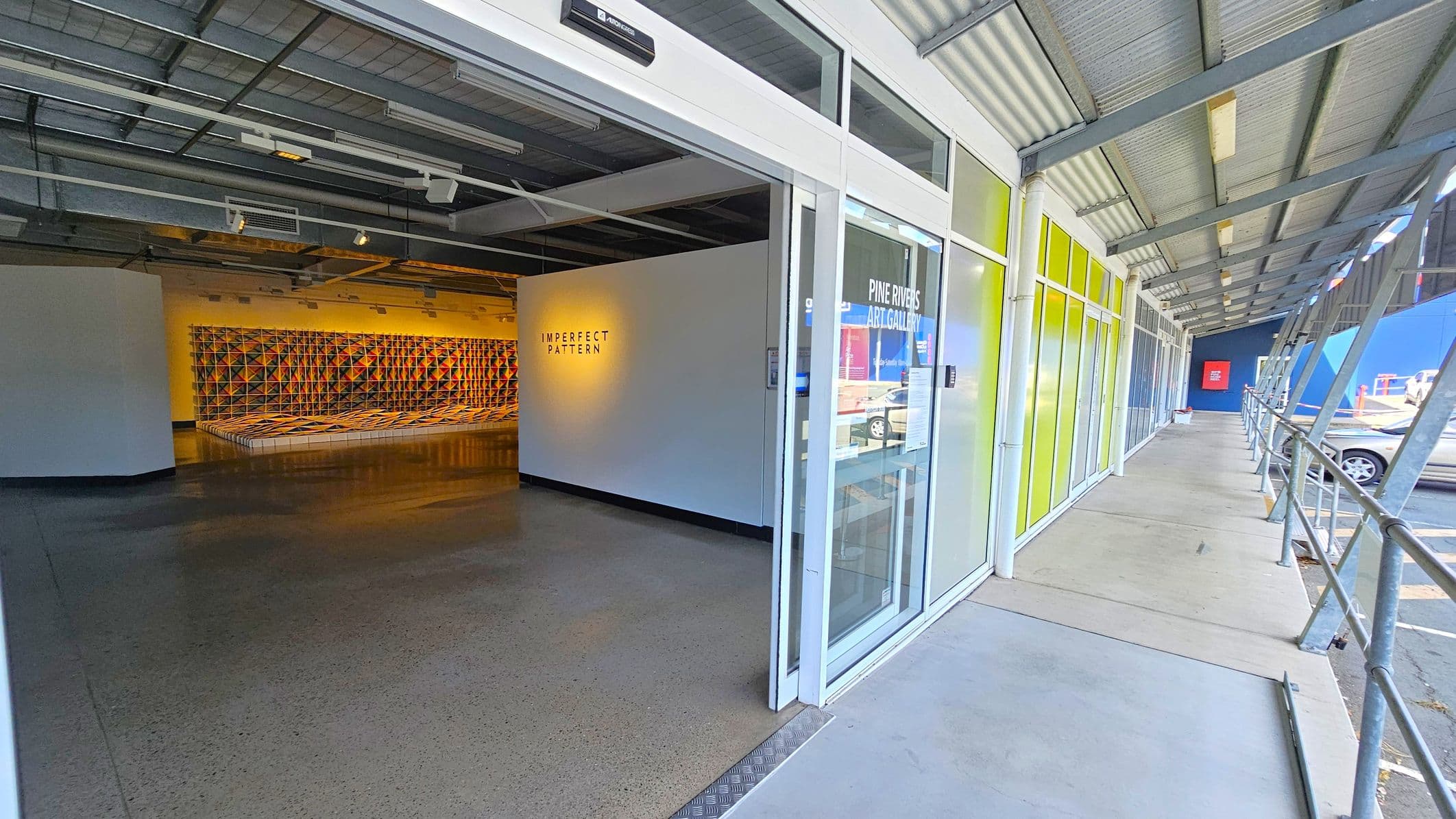 Entrance to an art gallery with a glass door on the right, labelled "Pine Rivers Art Gallery". The exterior features a covered walkway with a metal roof and railing. Inside, a wall displays the text "IMPERFECT PATTERN" with a geometric art installation in the background. The floor is polished concrete, and the ceiling has exposed beams and lighting fixtures.