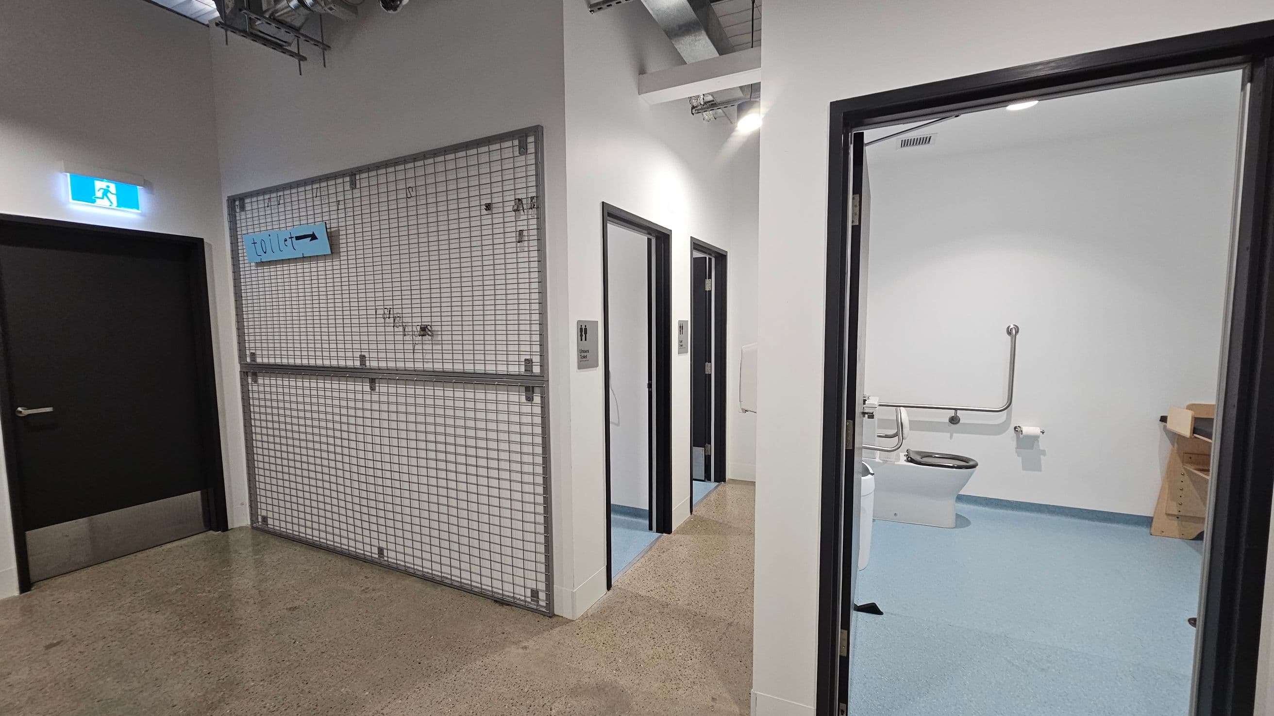 On the left, a black door with an illuminated exit sign above it. Next to it, a metal grid wall with a blue "toilet" sign. In the centre, two open doors with signs indicating male and female toilets. On the right, an open door reveals a bathroom with a toilet, grab bars, and a toilet roll holder. The floor is a mix of polished concrete and blue vinyl, and the ceiling has exposed ductwork.