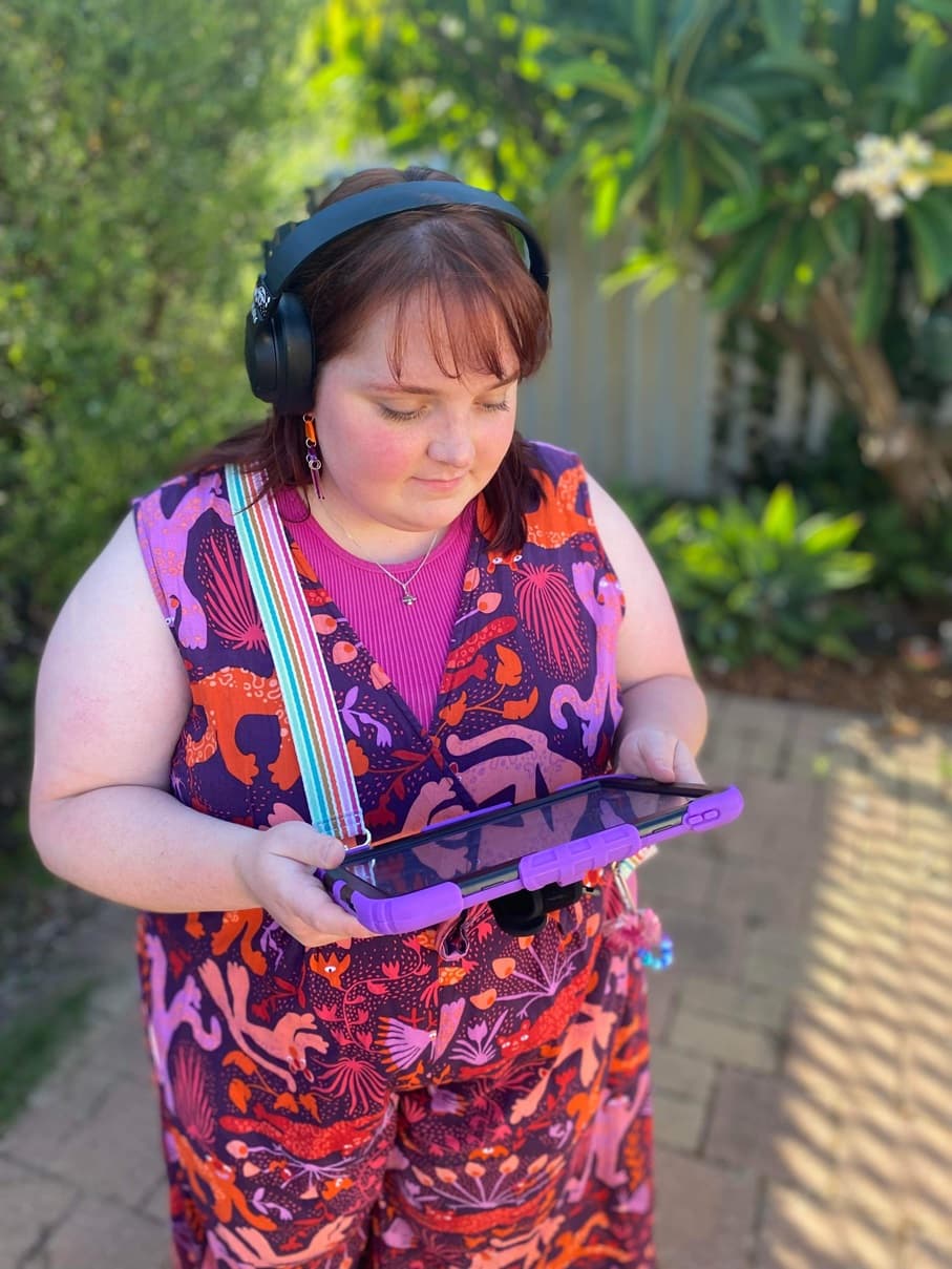 A woman wearing headphones and a colourful dress, looking at a tablet outdoors.